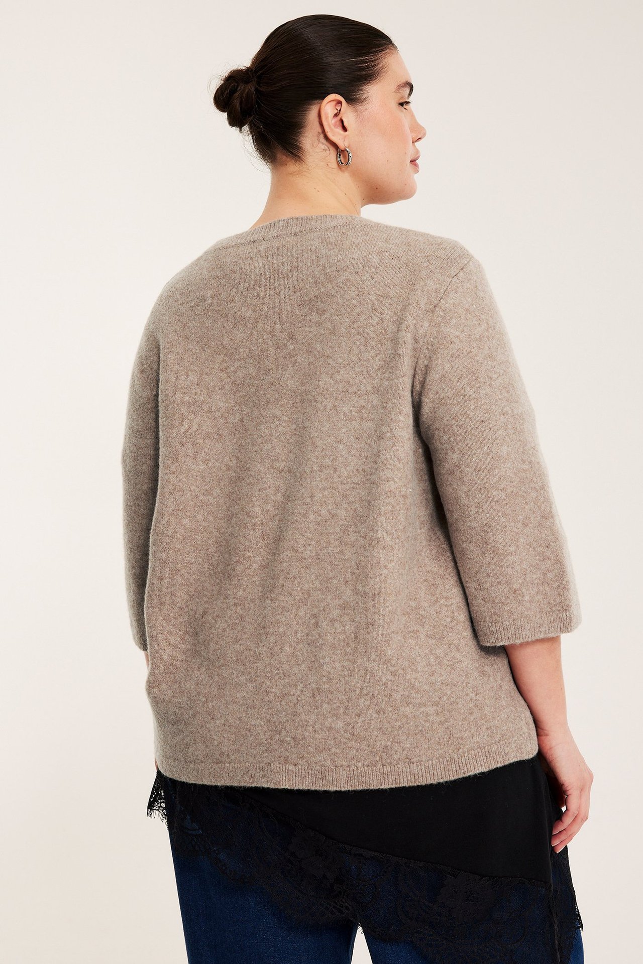 Naisen takakuva: ruskeamelange-neuletoppi 3/4-hihoilla, mustan pitsikoristeen ja sinisten farkkujen päällä.