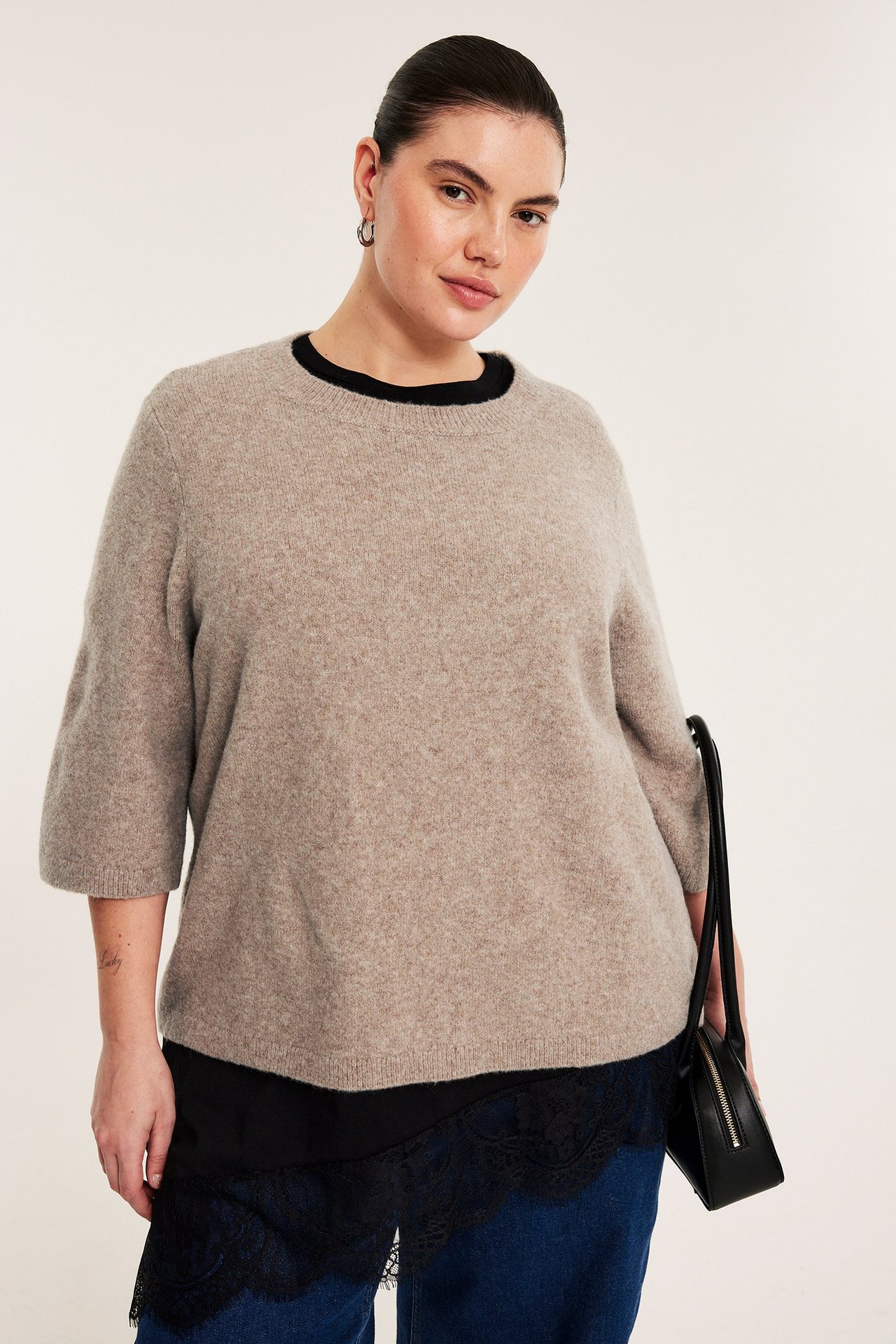 Nainen ruskeamelange 3/4-hihaisessa neulepaidassa, musta toppi, siniset farkut, musta käsilaukku.