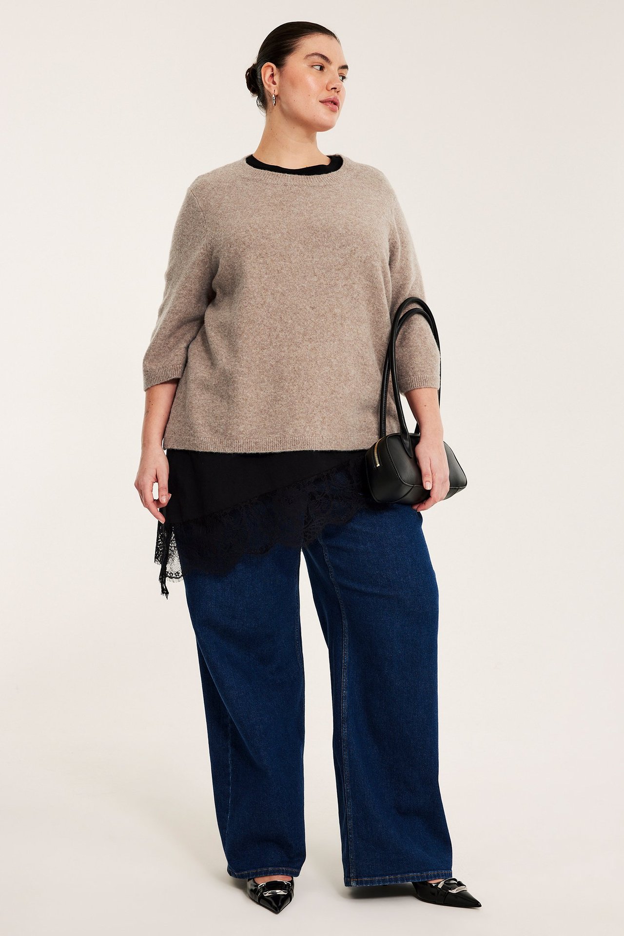 Edestä: Nainen meleratussa ruskeassa 3/4-hihaisessa neulepuserossa, musta pitsihelma, siniset farkut, musta käsilaukku.