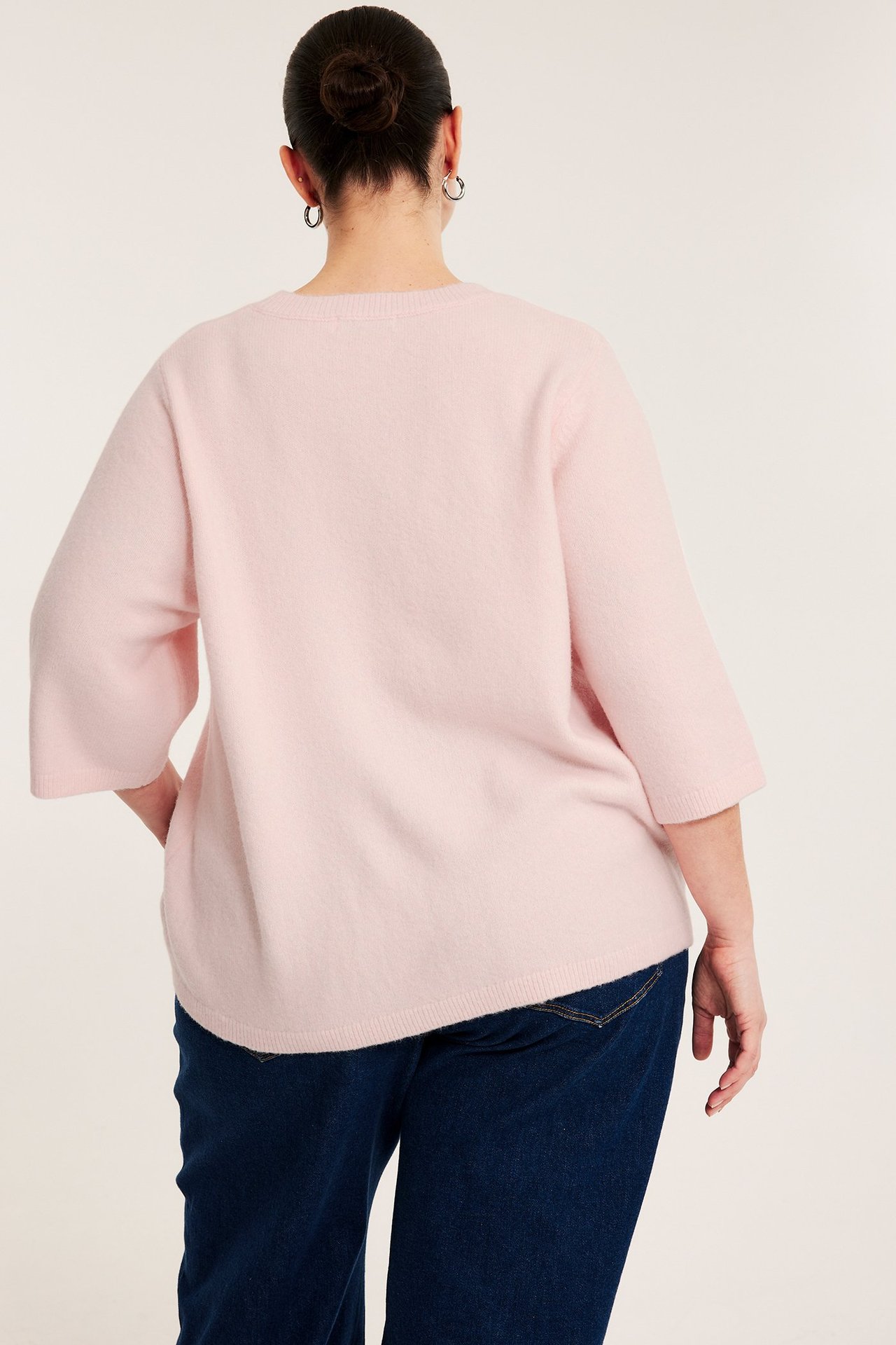 Bakifrån: Kvinna i ljusrosa stickad tröja med 3/4-ärm, blå jeans och silverfärgade ringörhängen.