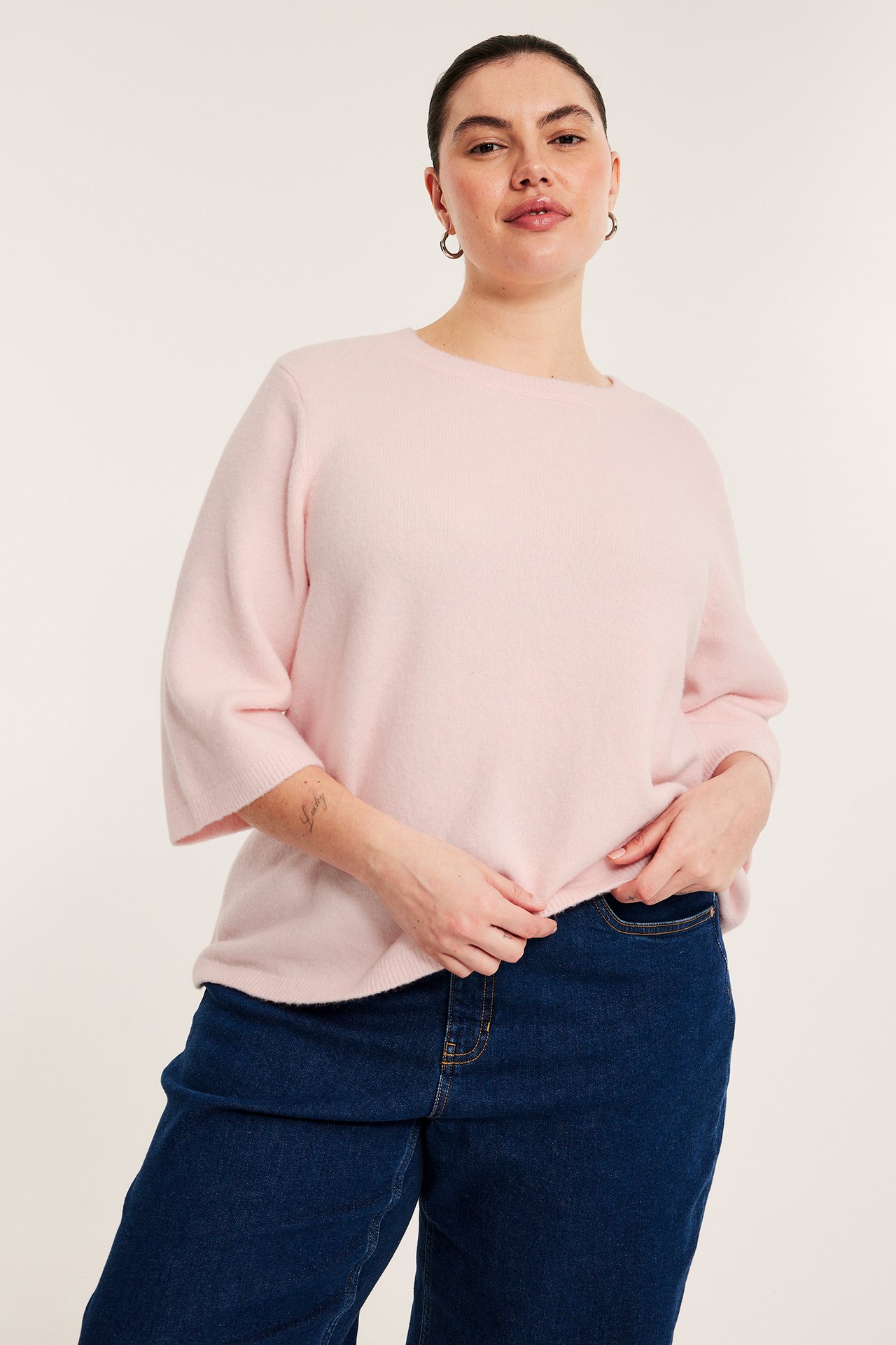 Frontvy av en kvinna i ljusrosa melerad stickad tröja med 3/4-ärm och mörkblå jeans.
