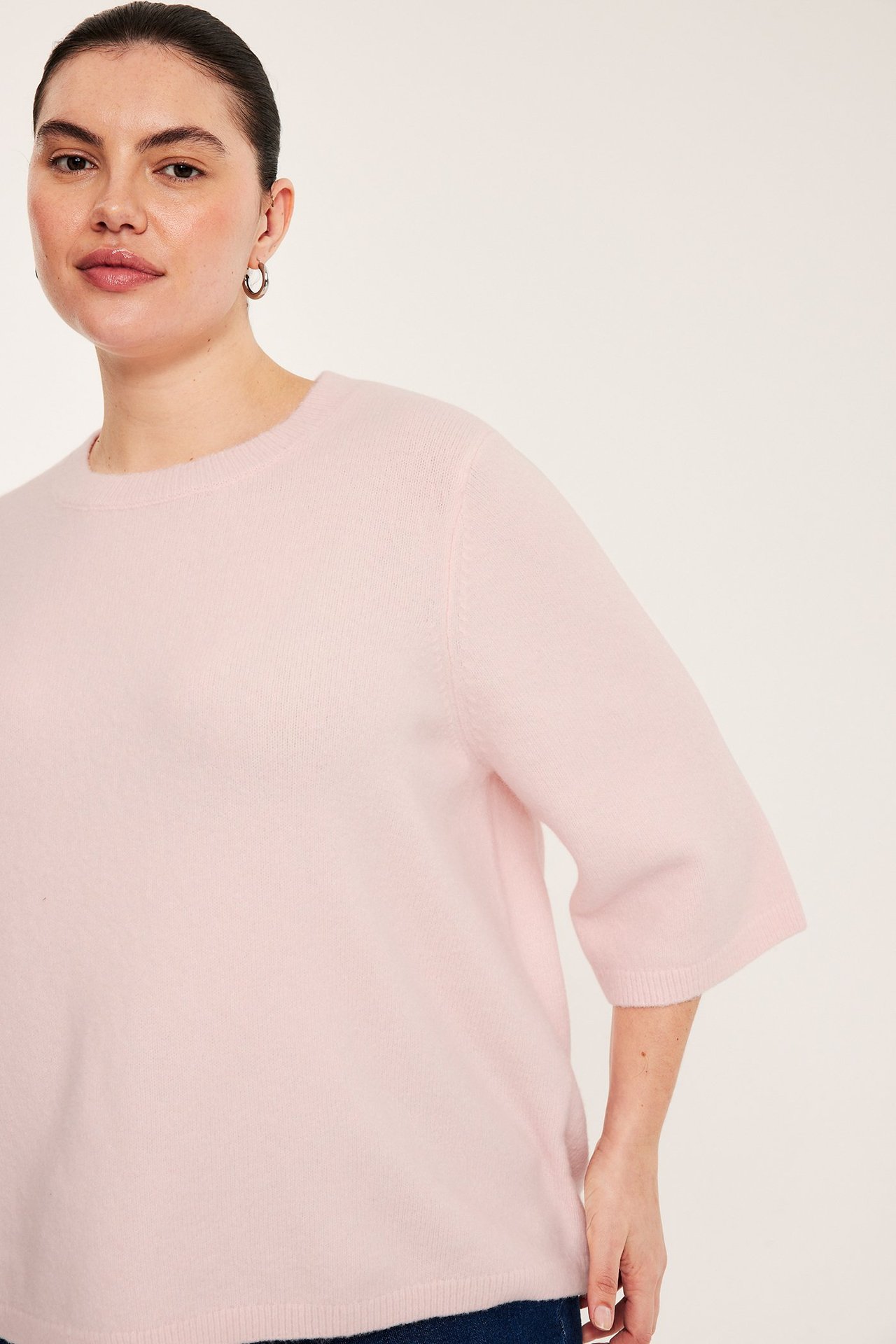 Framifrån: kvinna i ljusrosa melerad stickad topp, 3/4-ärm, rund halsringning och ringörhängen.