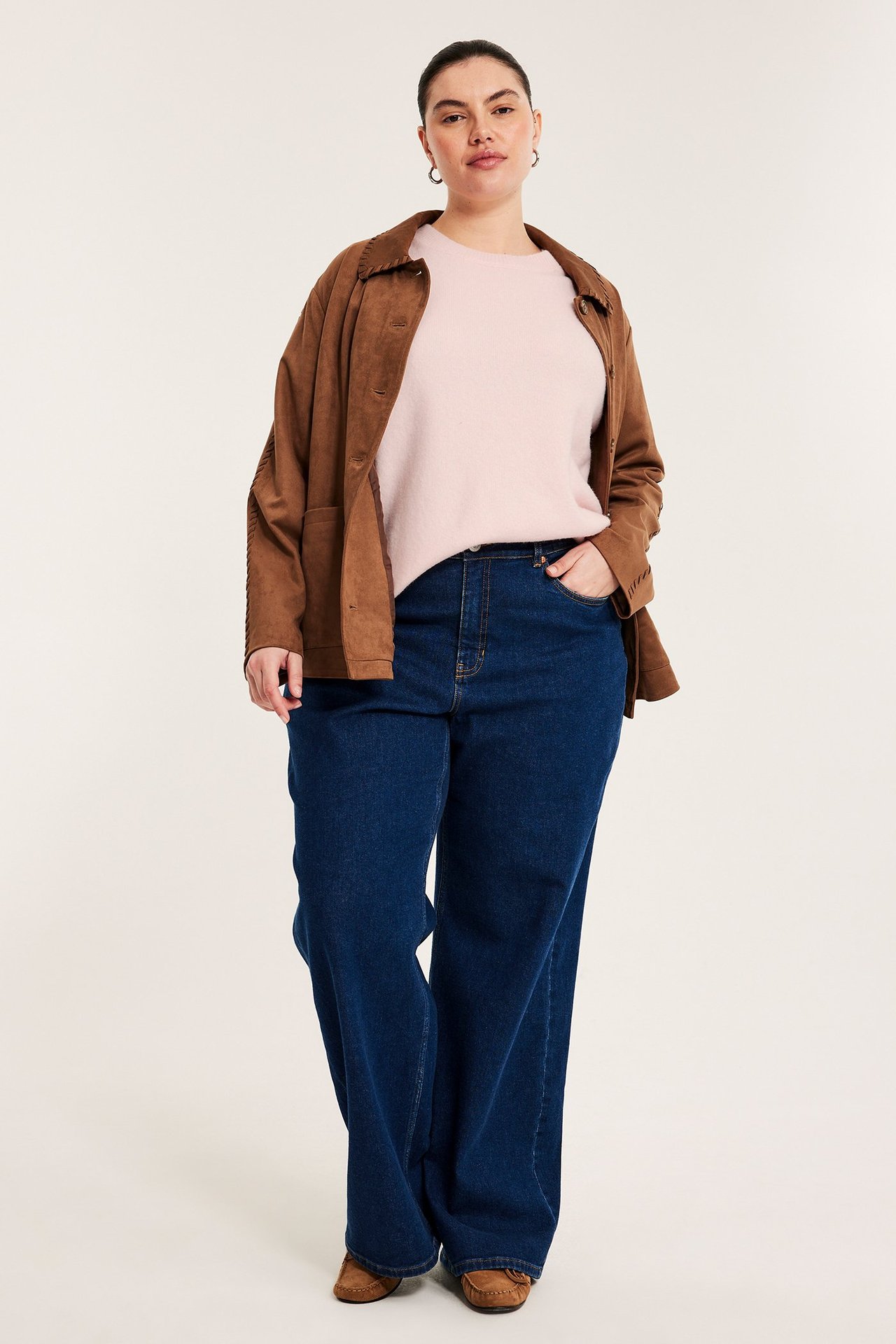 Framifrån, kvinna i ljusrosa melerad stickad topp, brun jacka och mörkblå vida jeans.