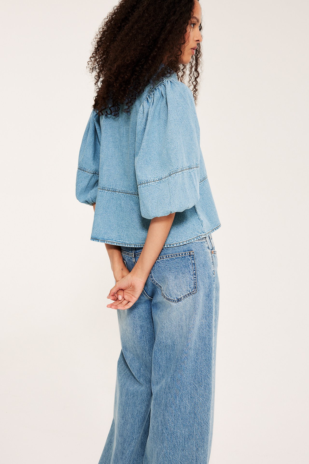 Kobieta od boku/tyłu: jasna dżinsowa bluzka peplum, bufiaste rękawy 3/4, niebieskie szerokie dżinsy.