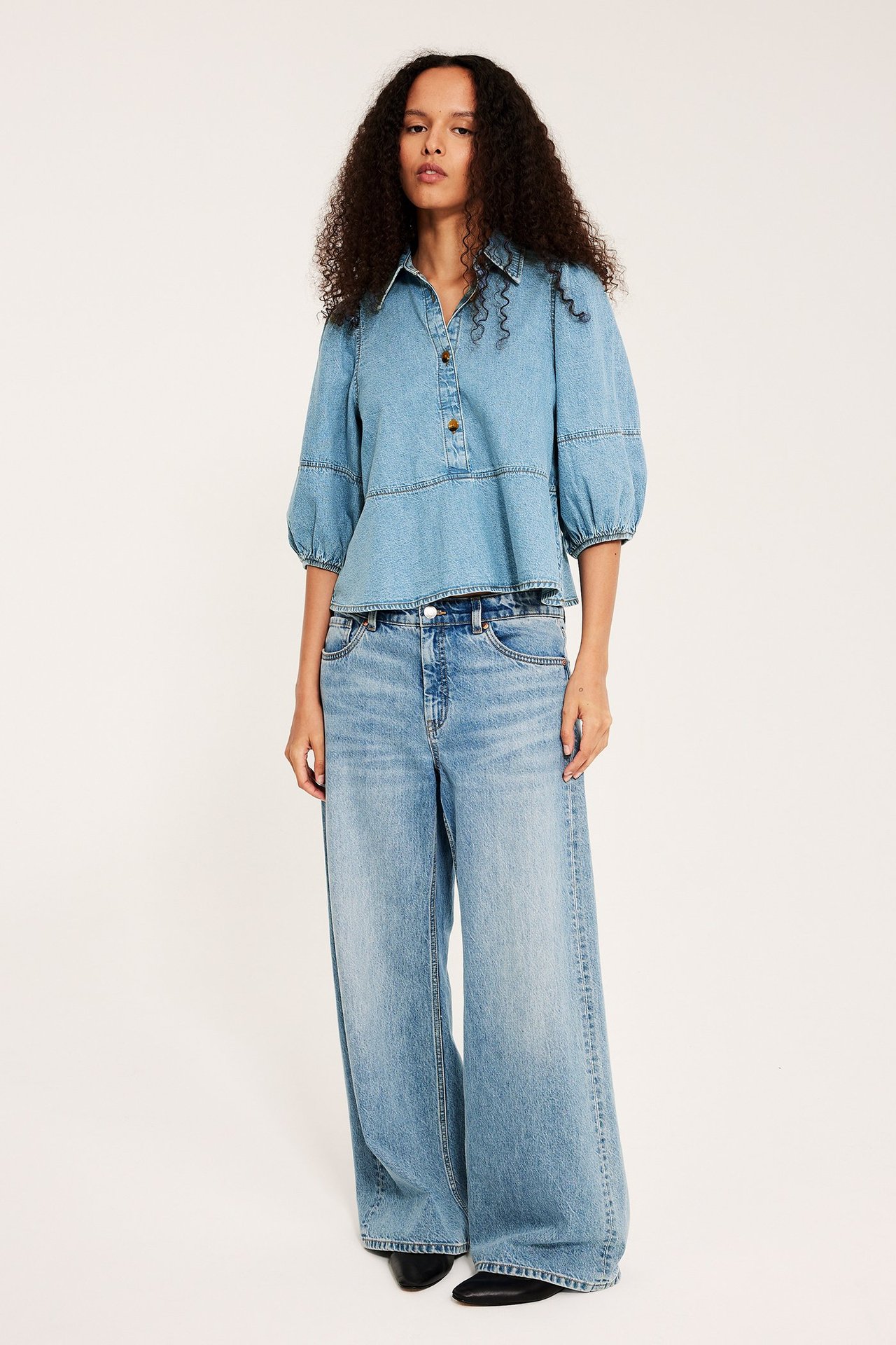 Nainen edestä: vaalea denimpeplum-pusero pussihihoilla, vaaleansiniset leveälahkeiset farkut, mustat kengät.