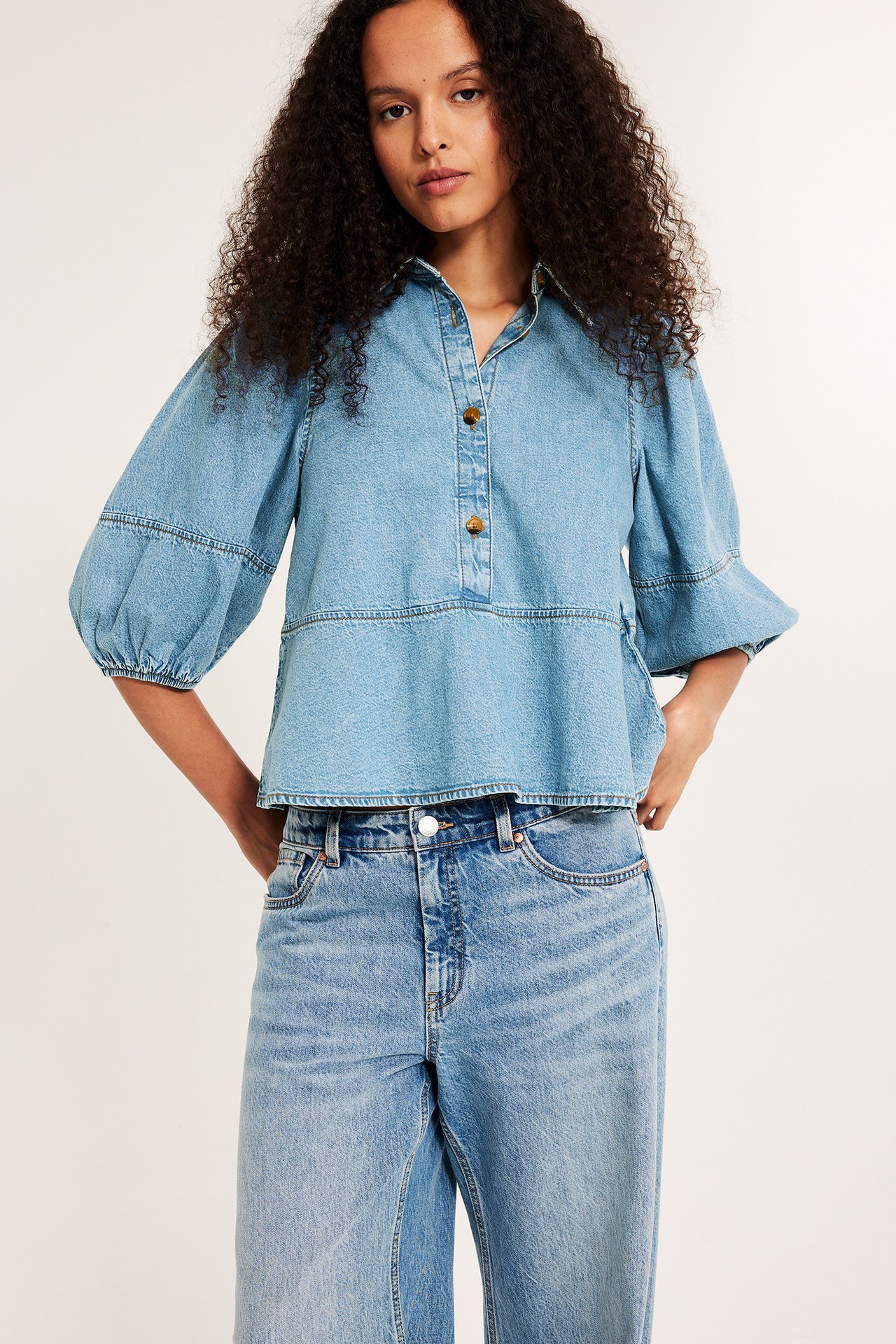Kobieta w jasnej jeansowej bluzce peplum z bufiastymi rękawami i rogowymi guzikami, z jeansami.