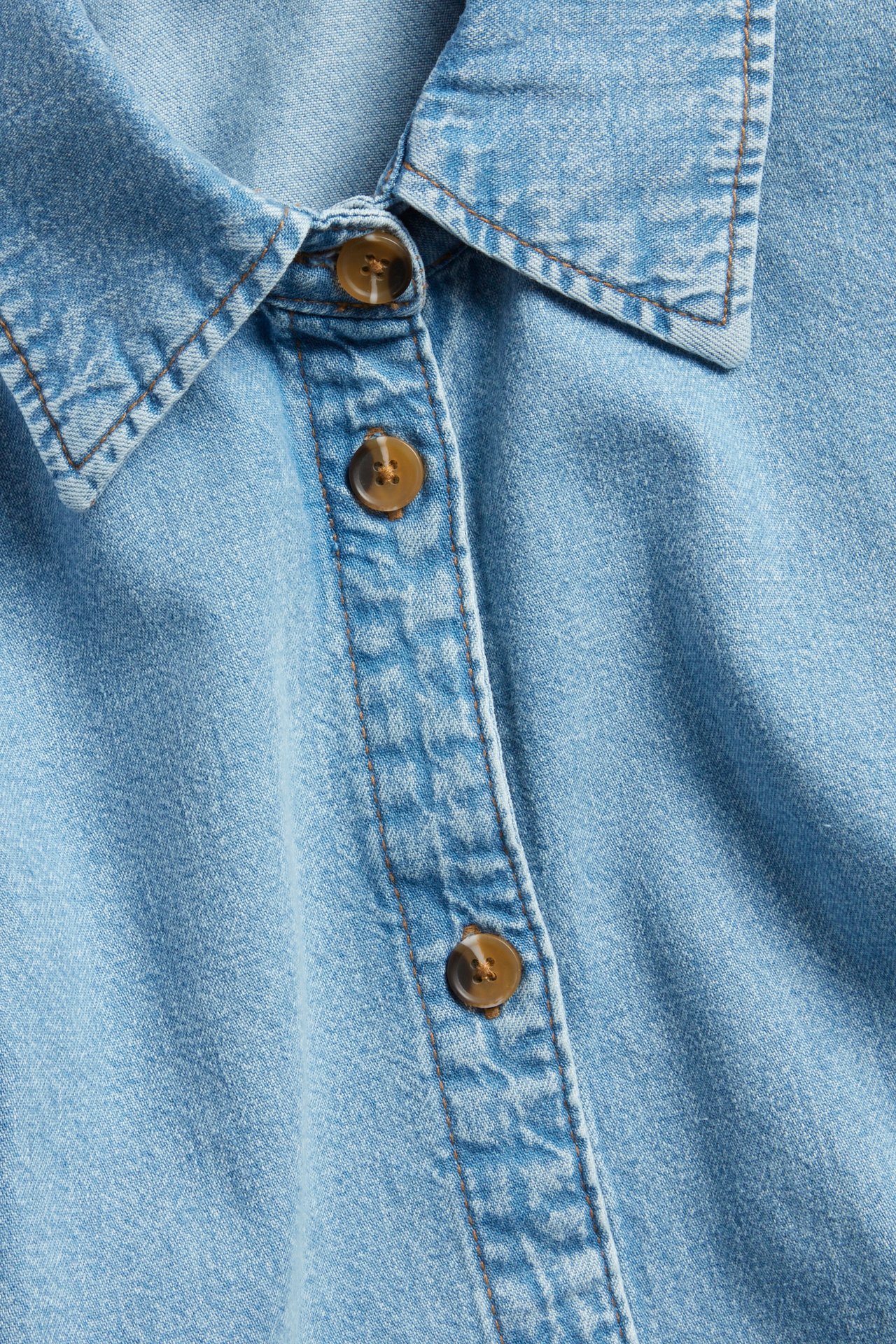 Lähikuva vaaleansinisestä denimbluusista, jossa on kaulus ja nappilista ruskeilla sarvinappikolla.
