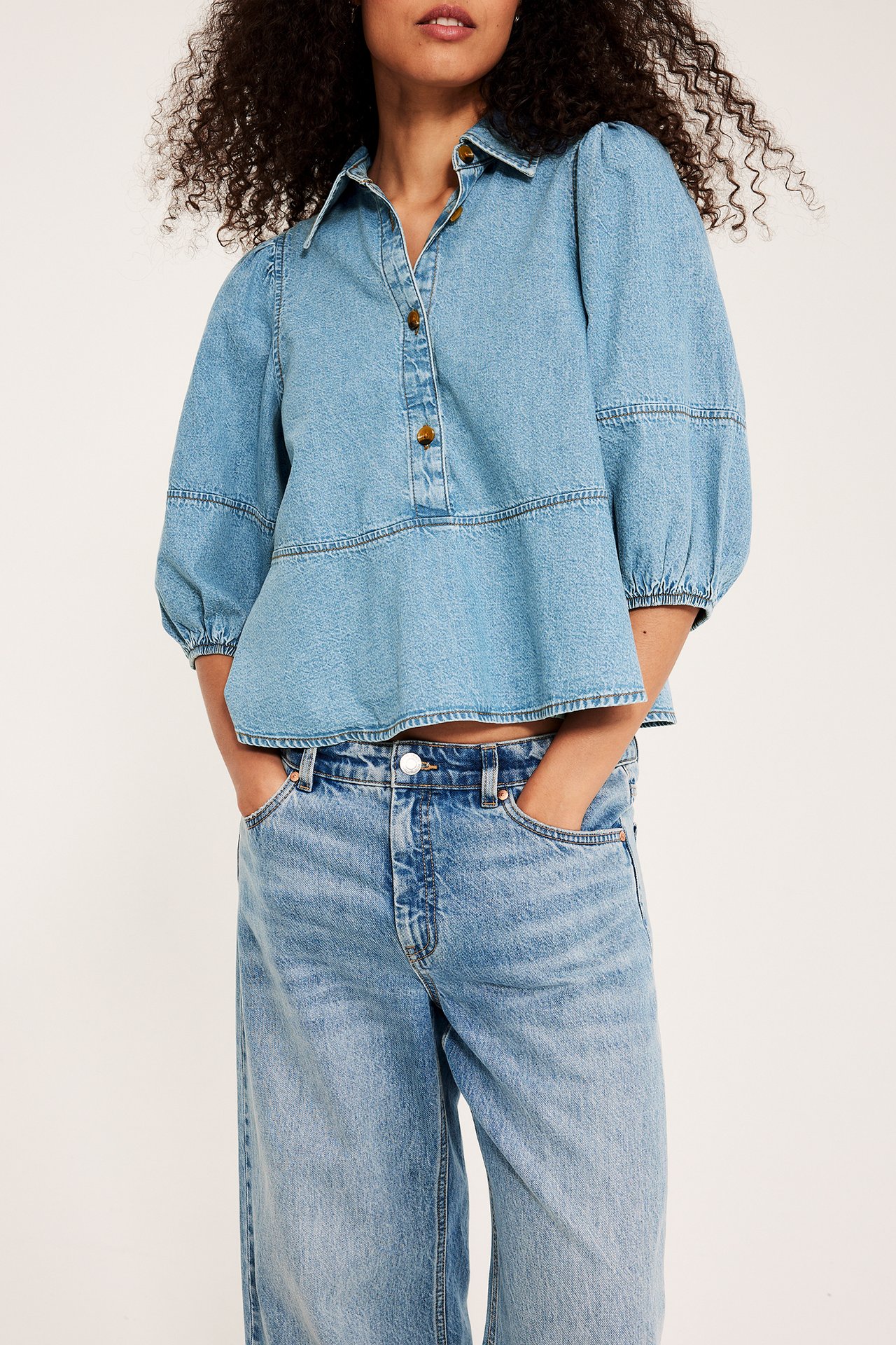 Widok z przodu: kobieta w jasnoniebieskiej bluzce peplum z guzikami rogowymi, bufiastymi rękawami i jeansach.