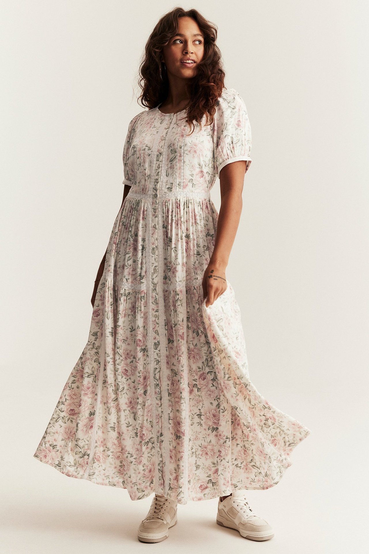 Widok z przodu: Kobieta w kwiecistej sukience maxi, z bufiastymi rękawami, koronką i beżowymi sneakersami.
