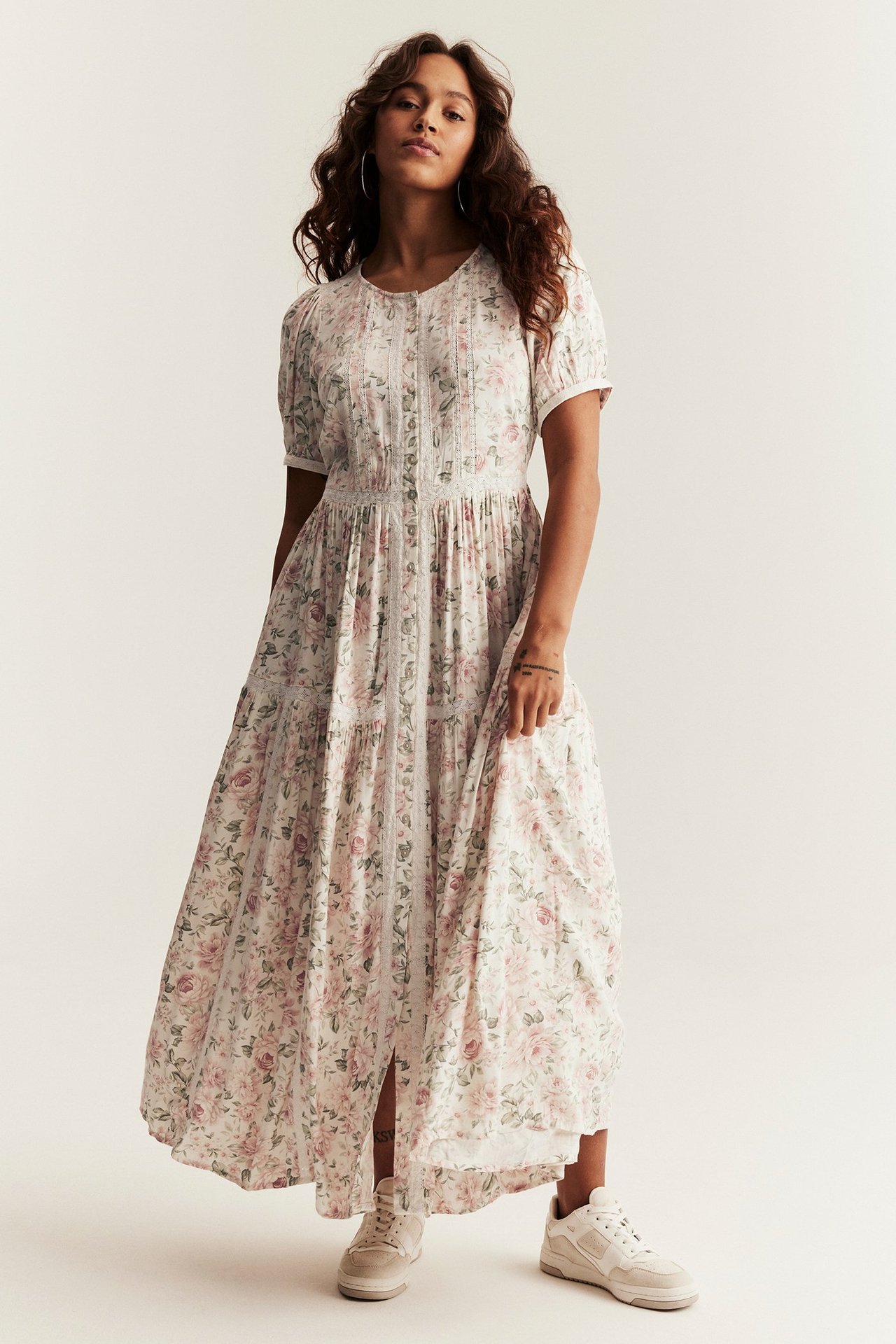 Front view: Kvinna i offwhite blommig maxiklänning med puffärm, spetsdetaljer, knappar fram, och beiga sneakers.