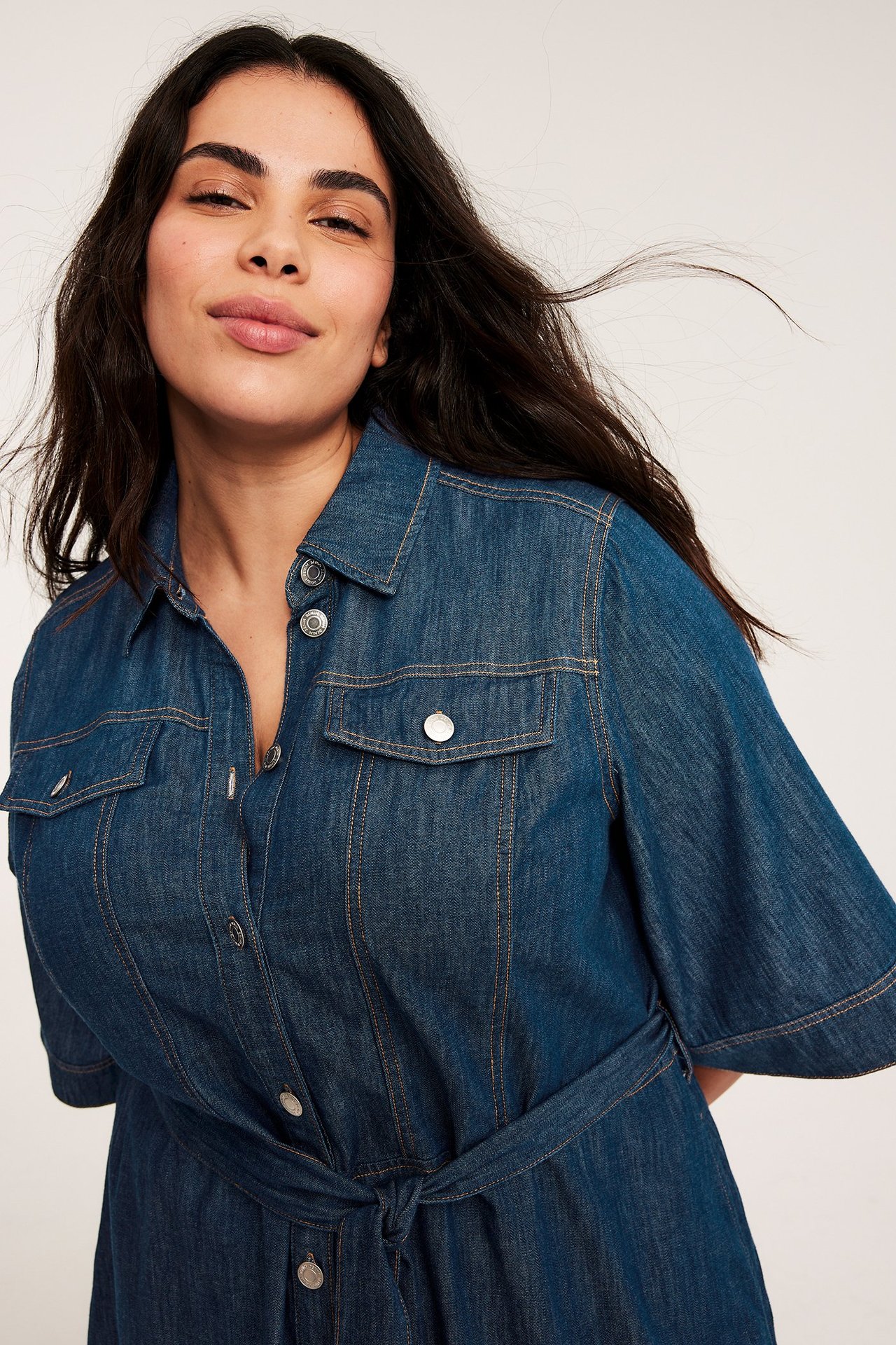 Närbild, framifrån, av en kvinna i en denim A-linjeklänning med knytband, korta ärmar och bröstfickor.