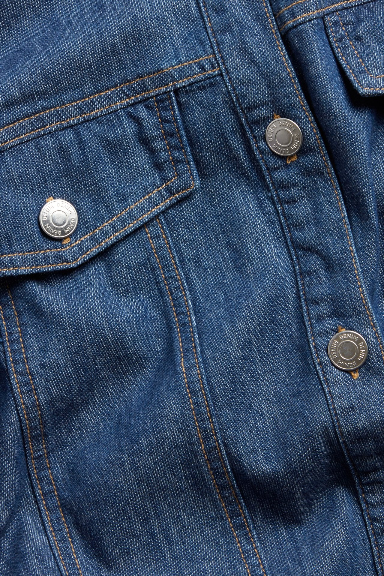 Lähikuva sinisestä farkkumekosta, jossa on hopeanväriset napit, ruskeat tikkaukset ja rintatasku.