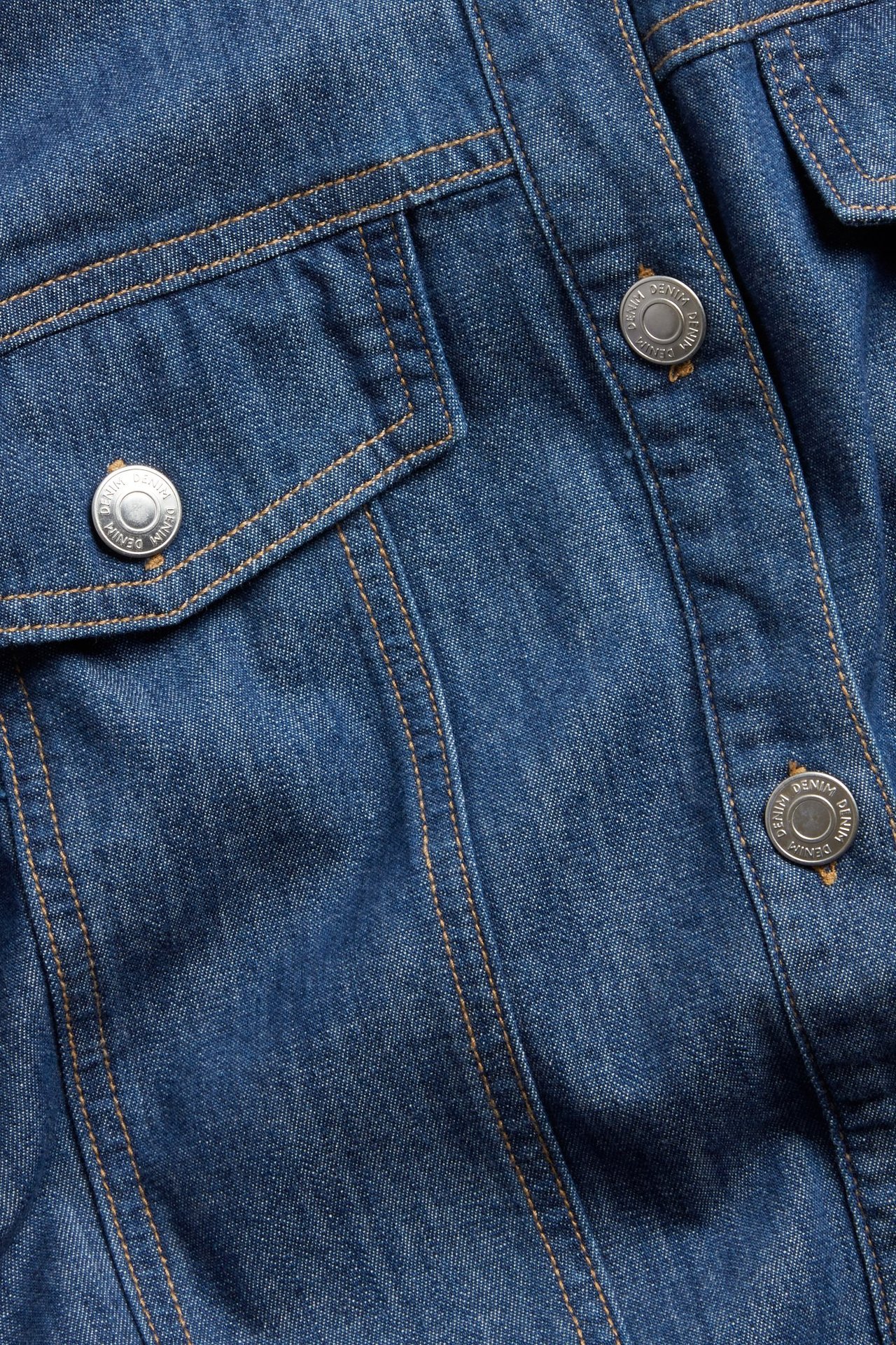 Zbliżenie na niebieską sukienkę jeansową z srebrnymi guzikami, brązowymi szwami i przednią kieszenią.