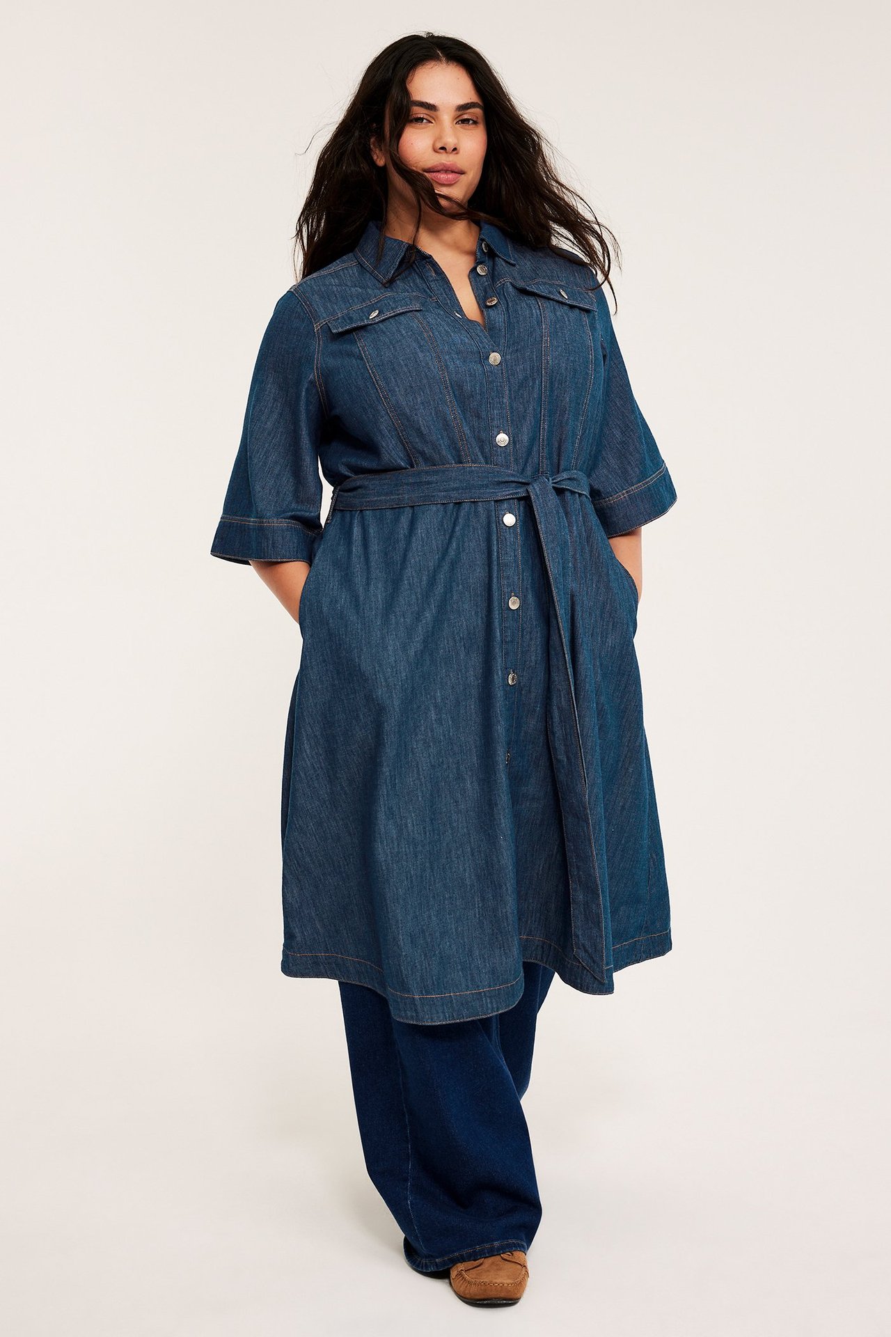 Framifrån: Kvinna i mörkblå A-linje jeansklänning med knytskärp, silverknappar, över vida jeans.