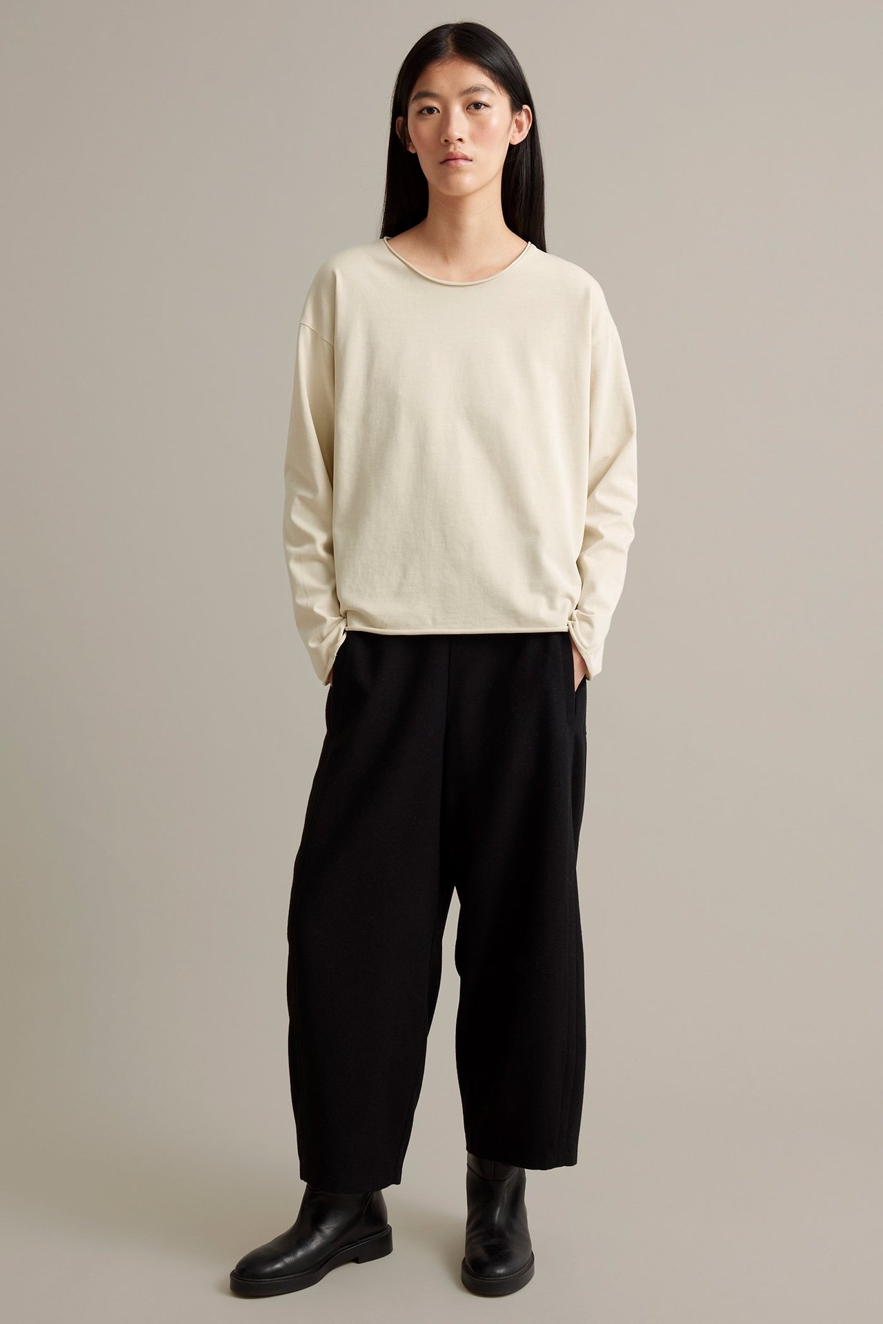 Nainen beigen pitkähihaisessa yläosassa ja tummissa löysissä housuissa, mustat bootsit, edestä.