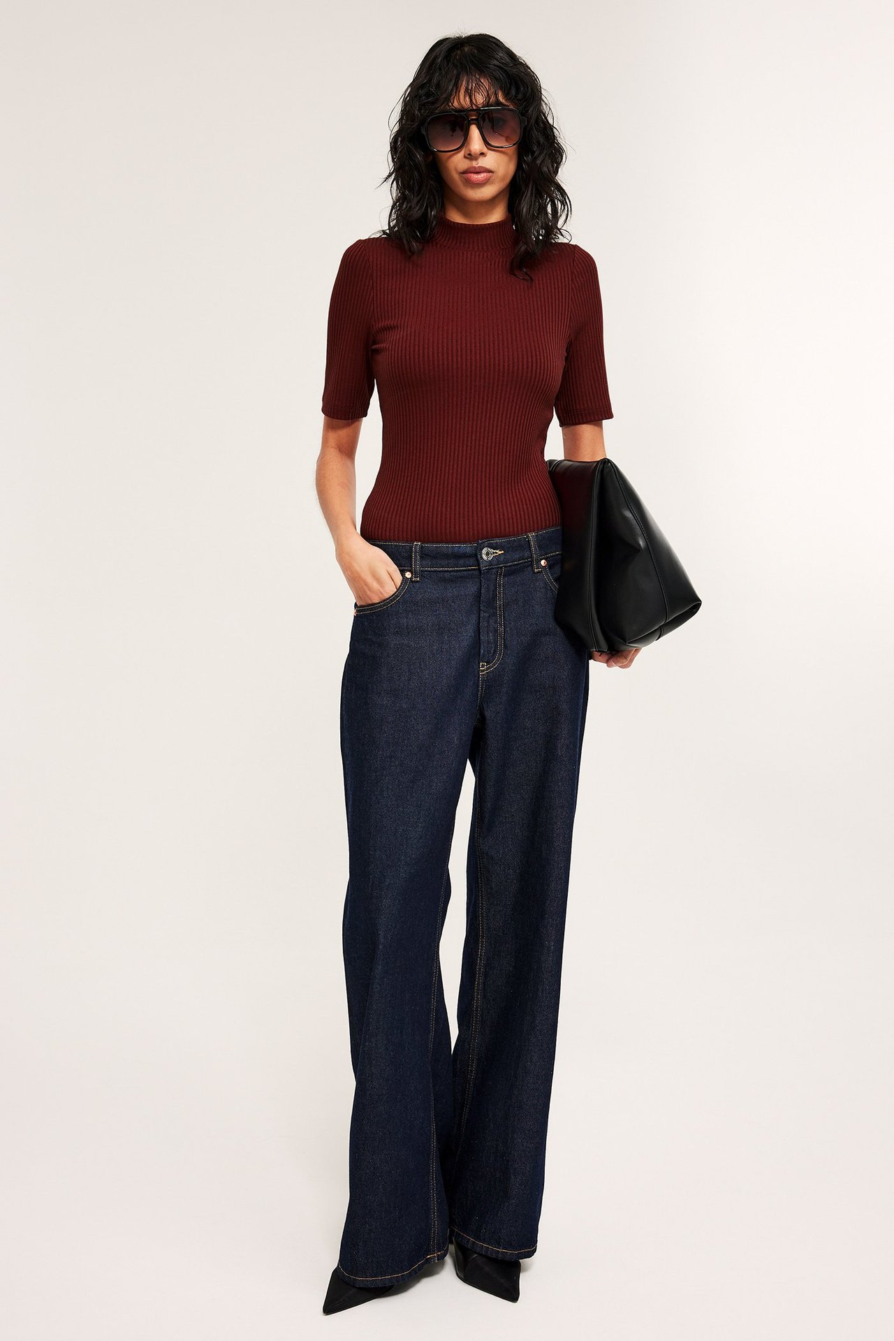 Kvinne i rød ribbestrikket polotopp, mørkeblå vide jeans, svarte hæler, solbriller, holder en svart veske.