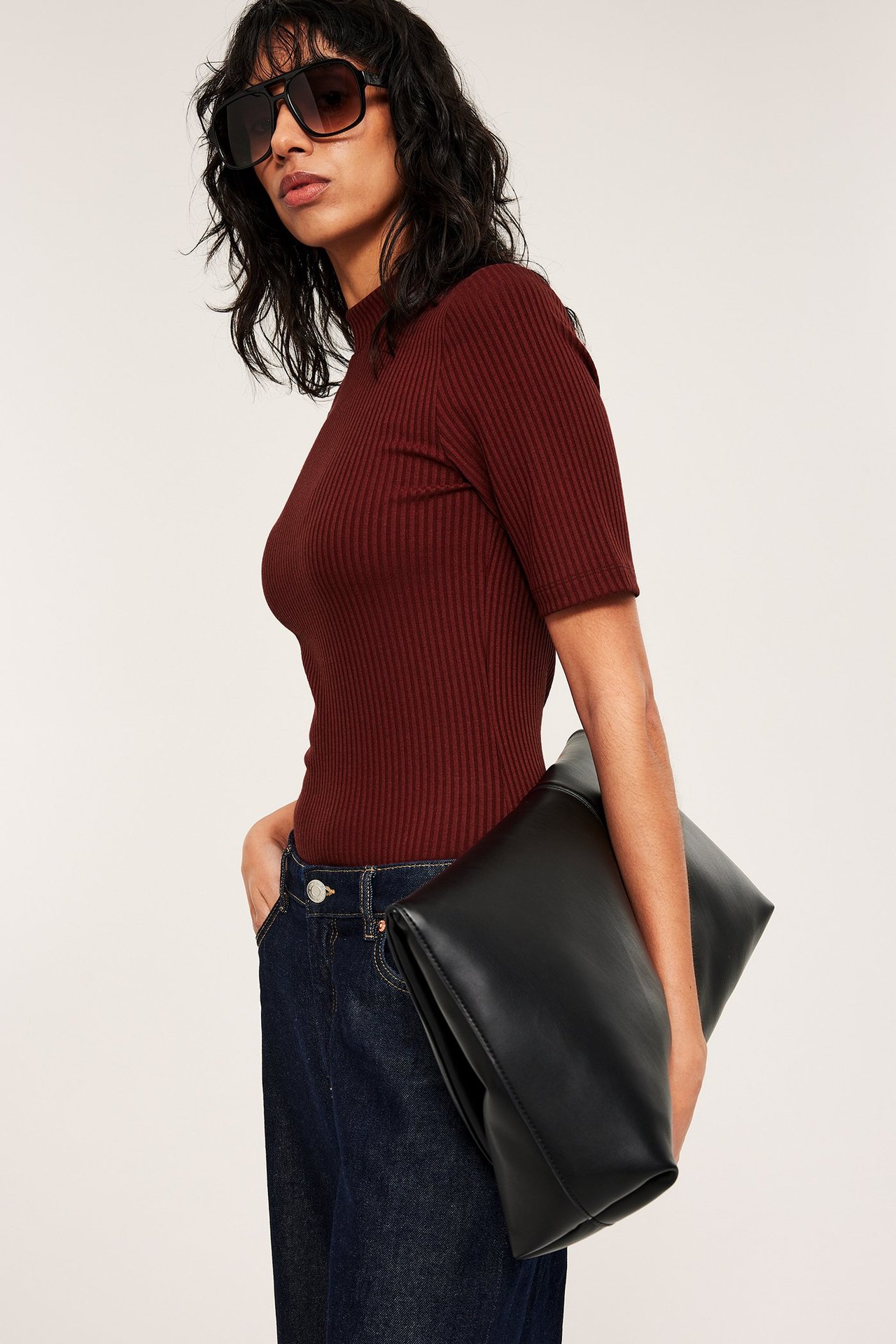 Sidebilde: Kvinne i mørkerød ribbet kortermet polotopp, mørkeblå jeans, svarte solbriller, holder en svart clutchveske.