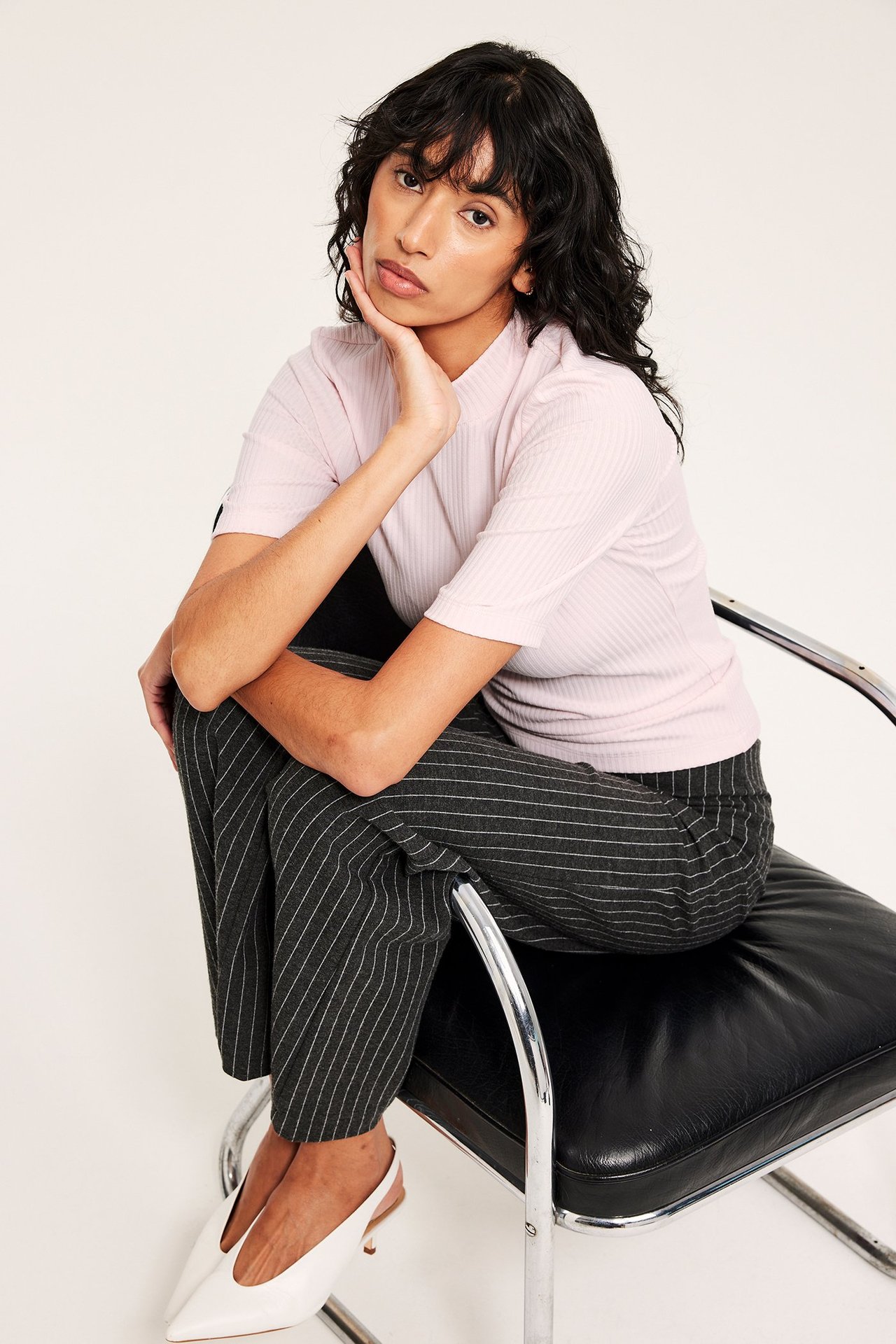 Kvinne i lyserosa ribbet kortermet polotopp, mørkegrå stripete bukser og hvite slingbacks.