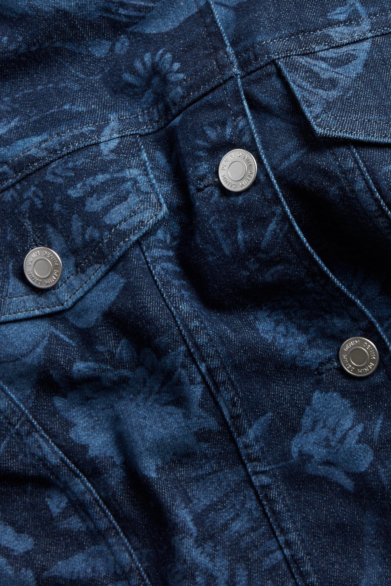 Zbliżenie na ciemnoniebieską kurtkę jeansową z subtelnym kwiatowym wzorem i metalowymi guzikami.