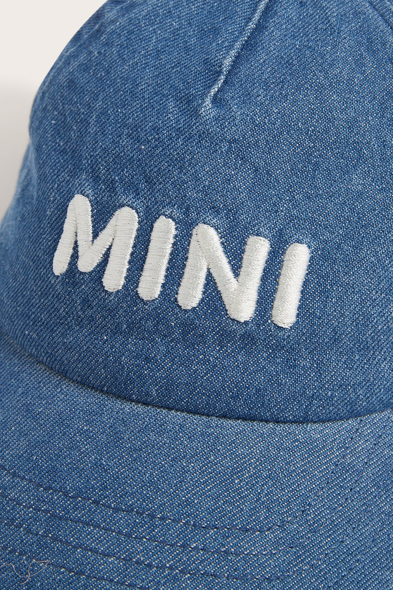 Zbliżenie od przodu na dżinsową czapkę z białym haftem 'MINI' z przodu.