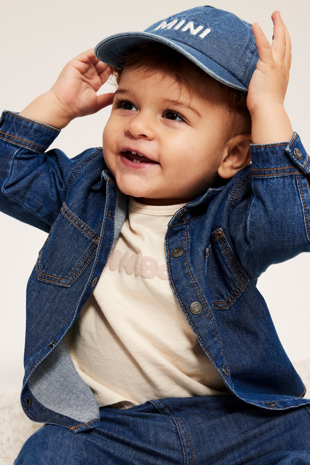 Baby har på seg en blå denimcaps med "MINI"-broderi, en denimskjortejakke, lys t-skjorte og denimbukser.