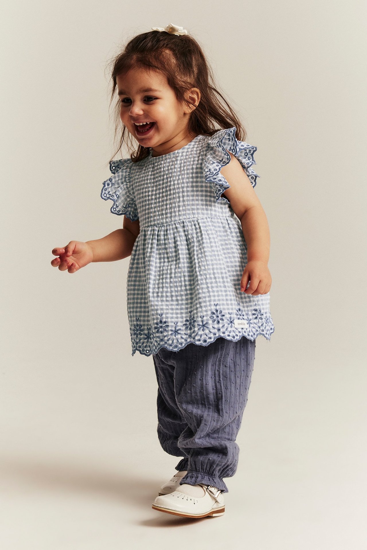 Forfra: En babyjente i en blå-hvitrutet blusebody med broderte volanger, blå mønstrede bukser, hvite sko.