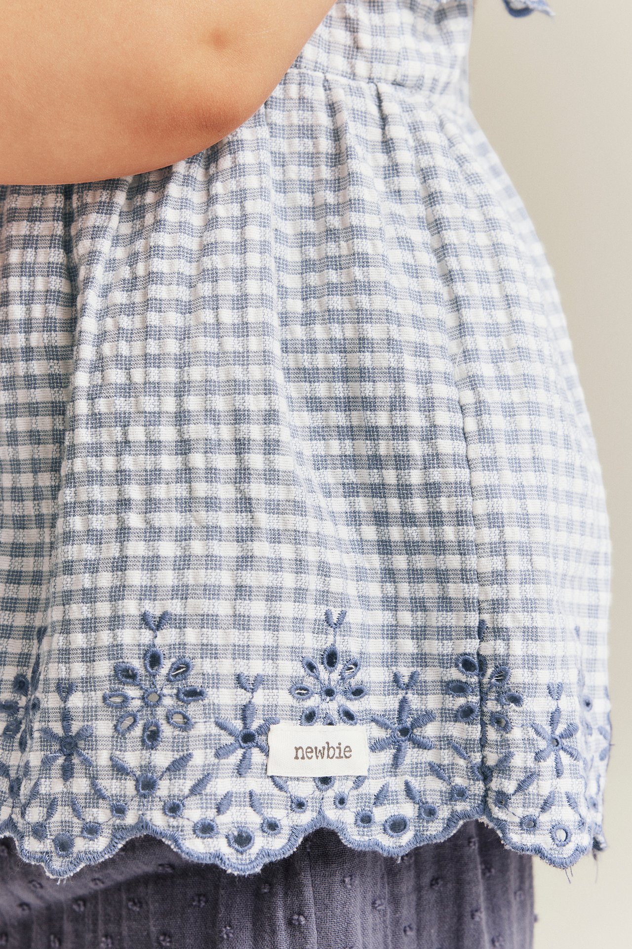Zbliżenie: niemowlęce niebiesko-białe w kratkę bawełniane body bluzkowe z baskinką i haftowanym falowanym brzegiem.