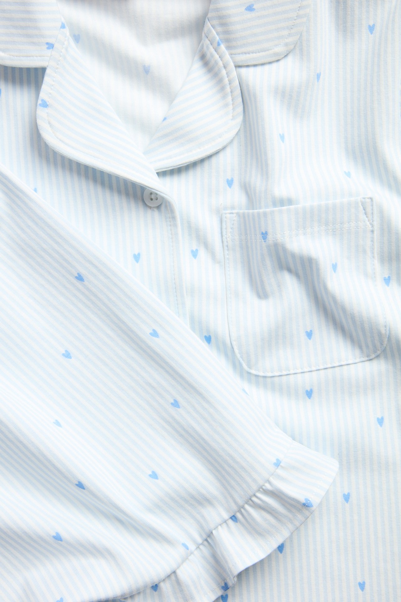 Närbild: Ljusblårandig pyjamastopp för barn med blå hjärtan, krage, knapp, ficka och volanger.