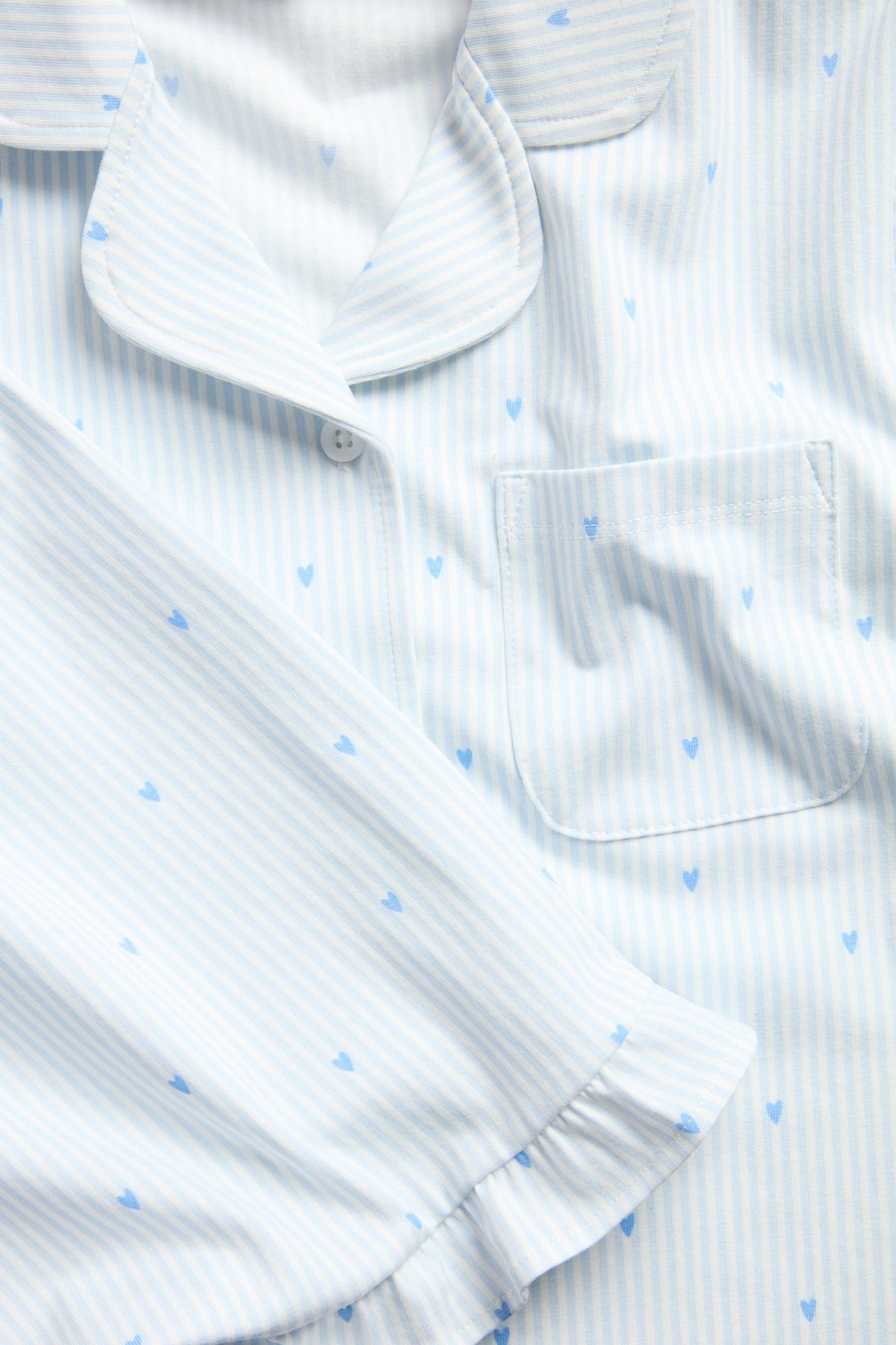 Zbliżenie: Dziecięca jasnoniebieska piżama w paski z niebieskimi sercami, kołnierzem, guzikiem, kieszenią i falbankami.