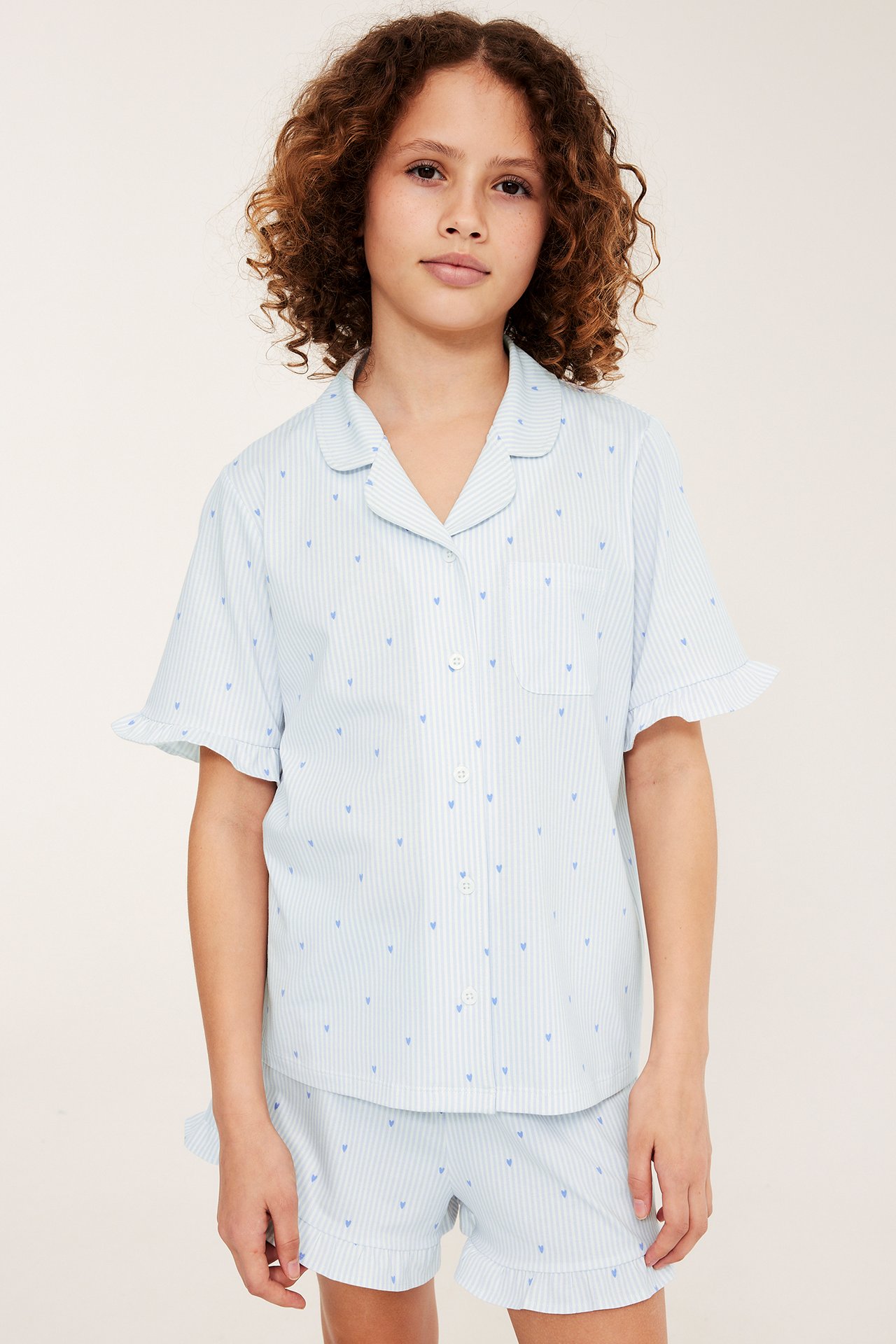 Frontutsikt av et barn i lyseblå stripete bomullspyjamas med blå hjerter, volangermer og matchende shorts.