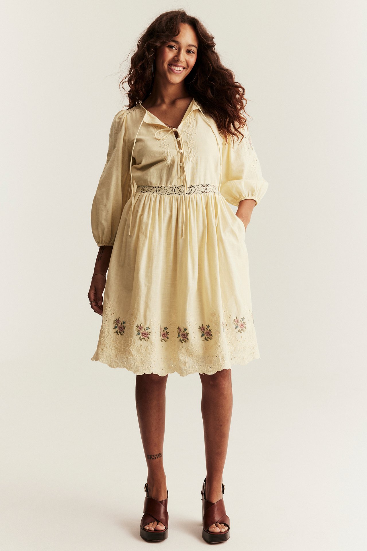 Frontvy av en leende kvinna i kort gul broderad klänning med puffärm och bruna platåsandaler.
