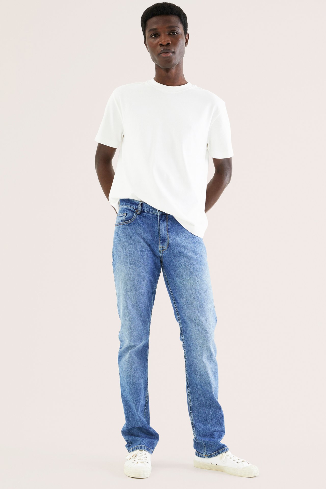 Framifrån, en man i ljusblå raka jeans, en vit rundhalsad t-shirt och vita sneakers.