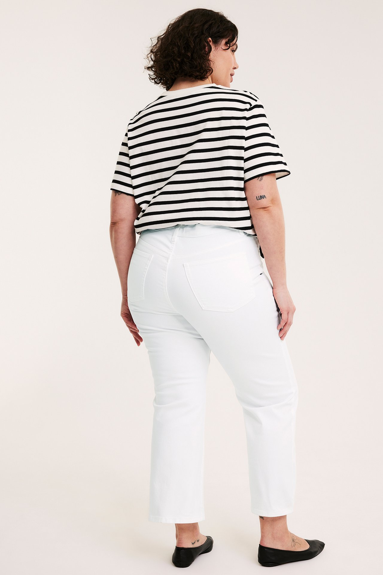 Ryggbilde av kvinne i hvite høylivede cropped flare-jeans, en stripete t-skjorte og svarte ballerinasko.