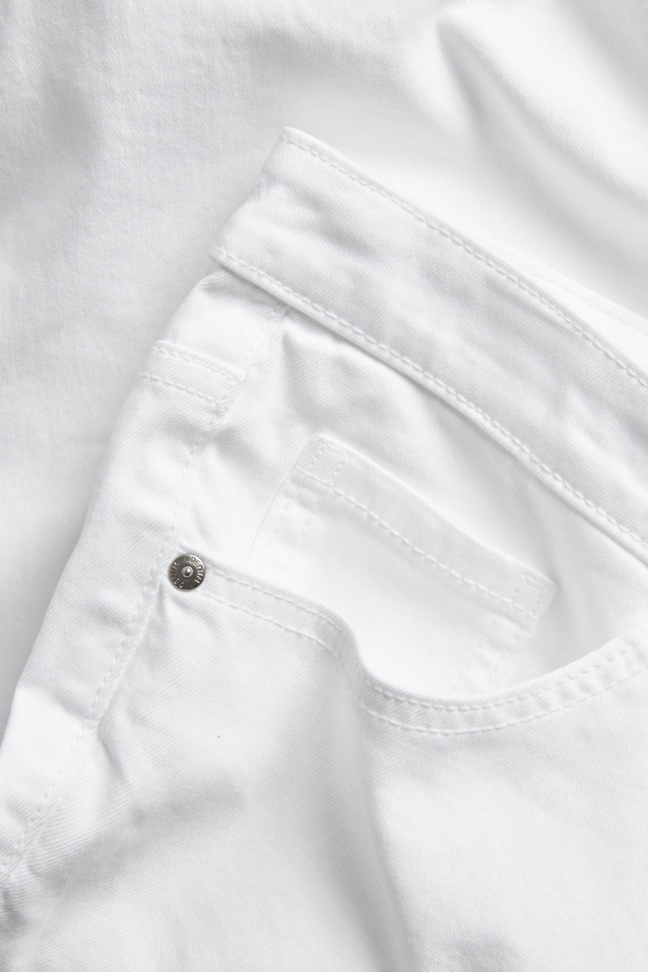 Nærbilde av hvite cropped flare-jeans, viser frontlommen med en sølvfarget metallnagle og sømmer.