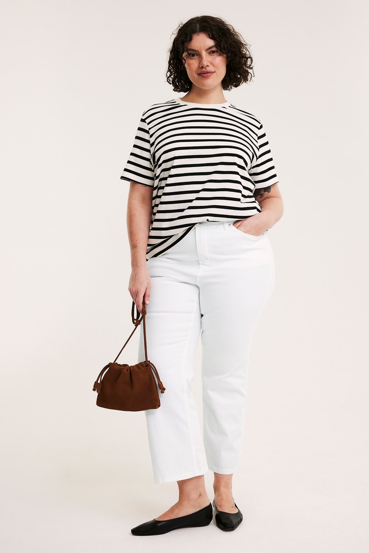 Frontvisning: Kvinne i hvite høylivs cropped flare-jeans, stripet topp, svarte flats, holder brun veske.