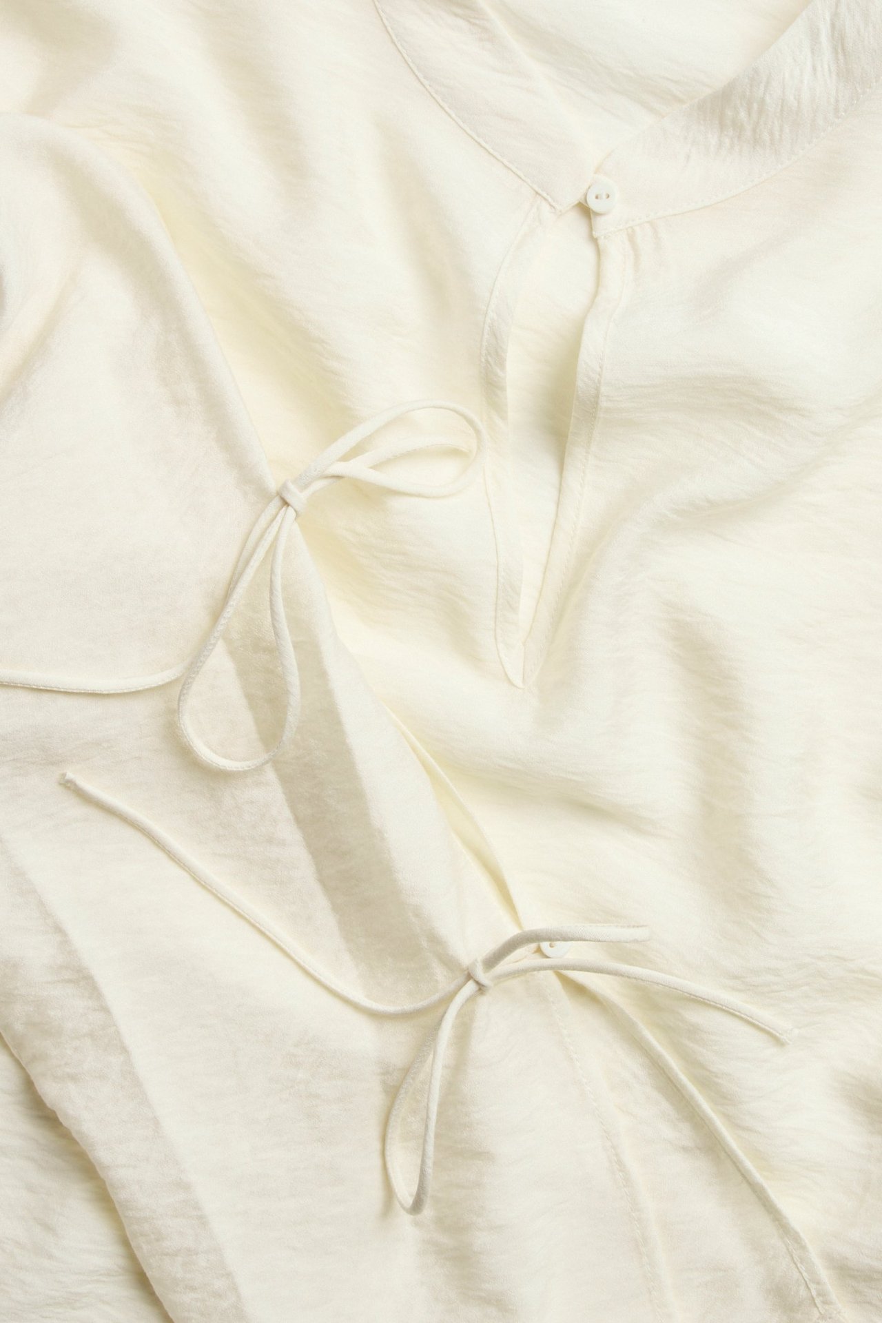 Zbliżenie na bluzkę w kolorze ecru z jednym guzikiem przy dekolcie i dwoma widocznymi wiązaniami.