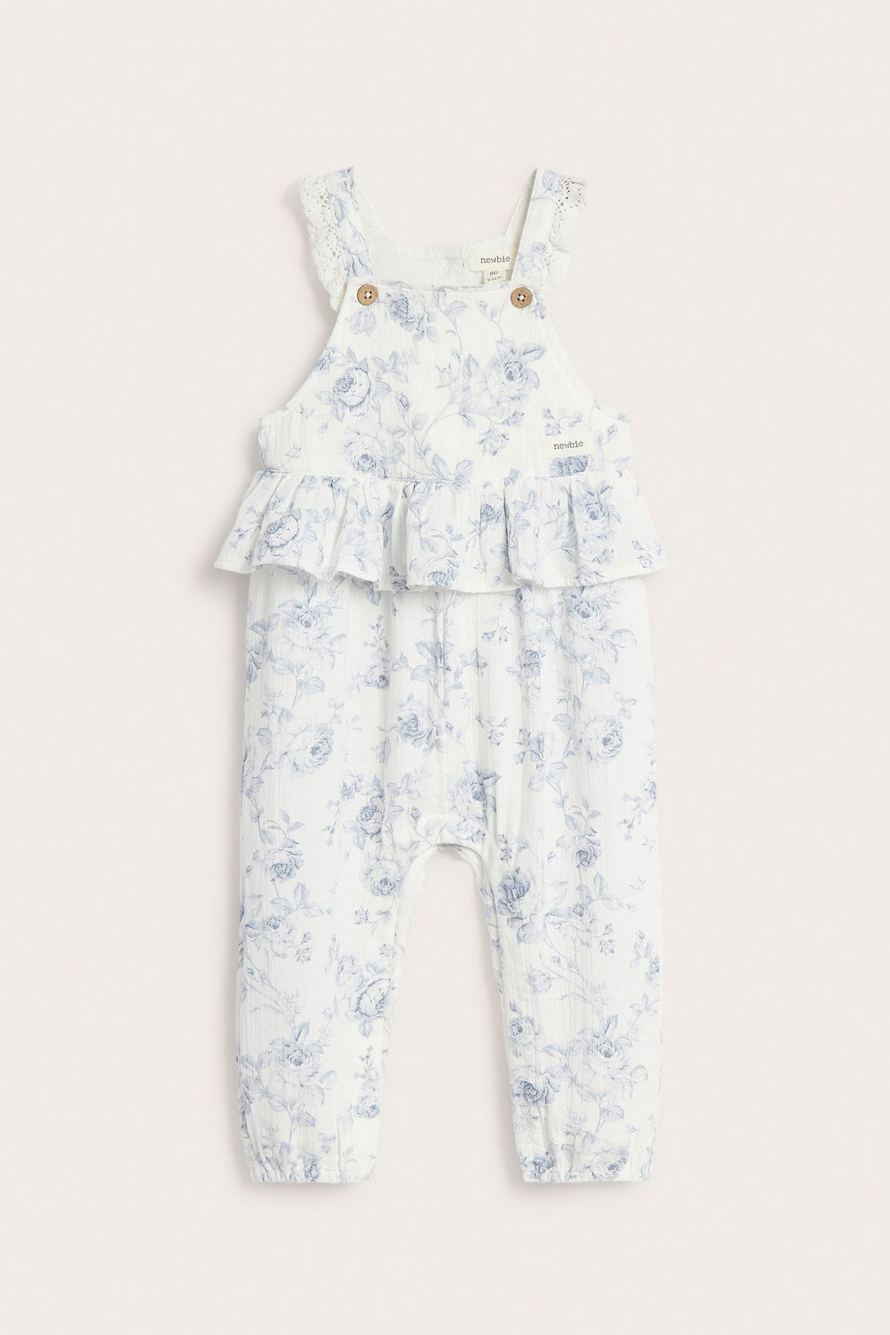 Forfra: Off-white baby-overall med blomstermønster, volang i midjen, blondekant og knappe-stropper.