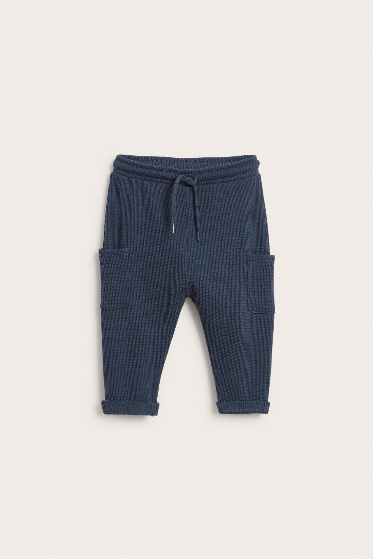 Frontvisning av mørkeblå, vaffelstrikkede babybukser med snøring i livet, sidelommer og oppbrettede kanter.
