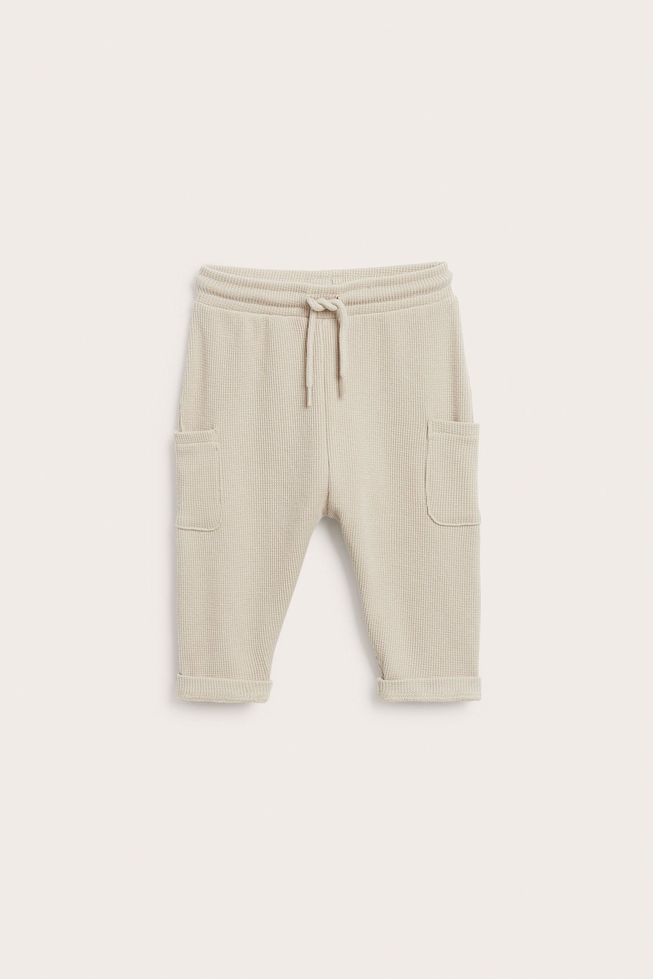 Sett forfra: Beige vaffelvevde babybukser med snøring i midjen, sidelommer og oppbrettede kanter.