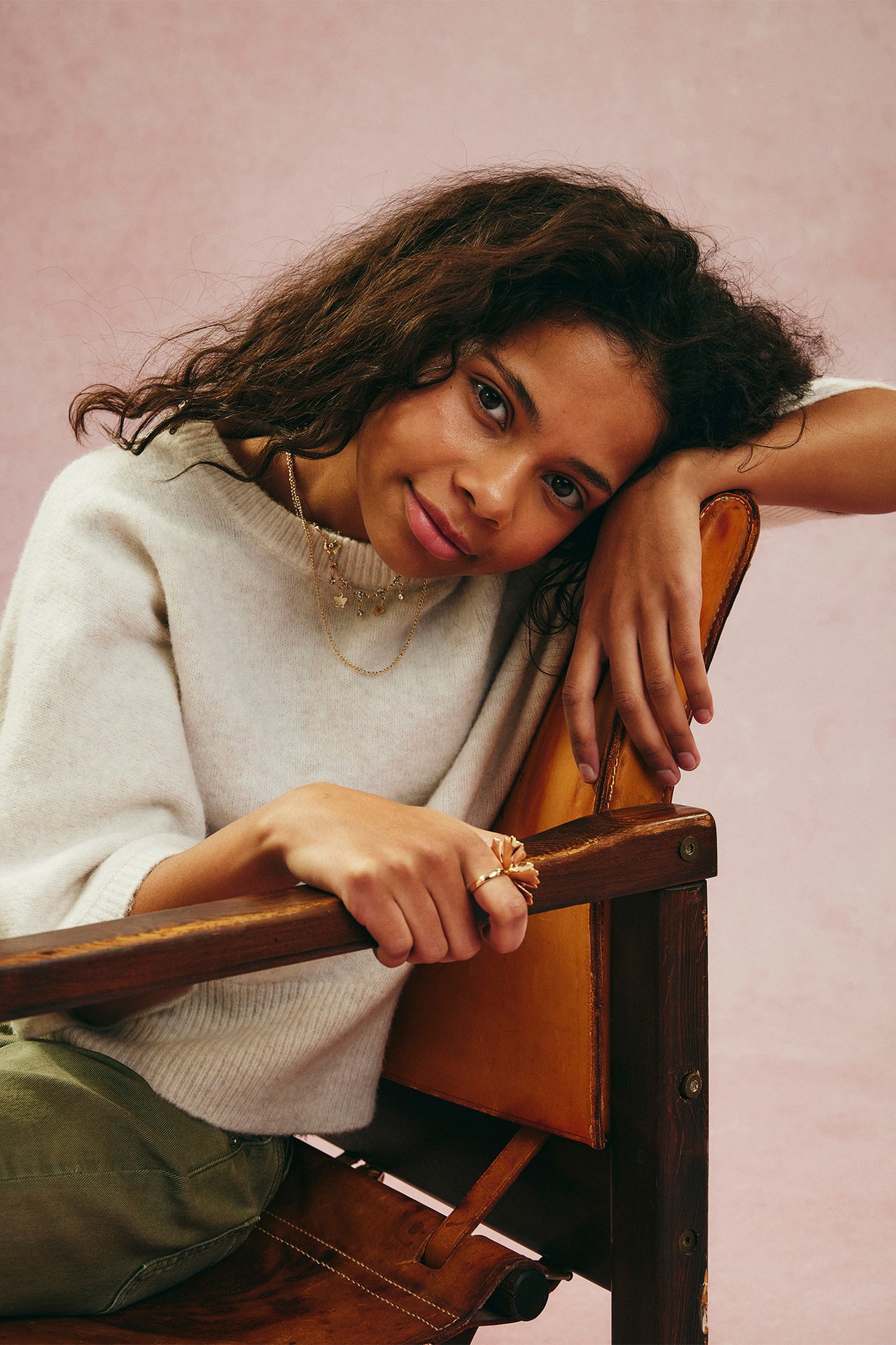 Widok z przodu: młoda kobieta w melanżowym beżowym dzianinowym topie, zielonych spodniach, złotym naszyjniku i złotym pierścionku z kwiatkiem.