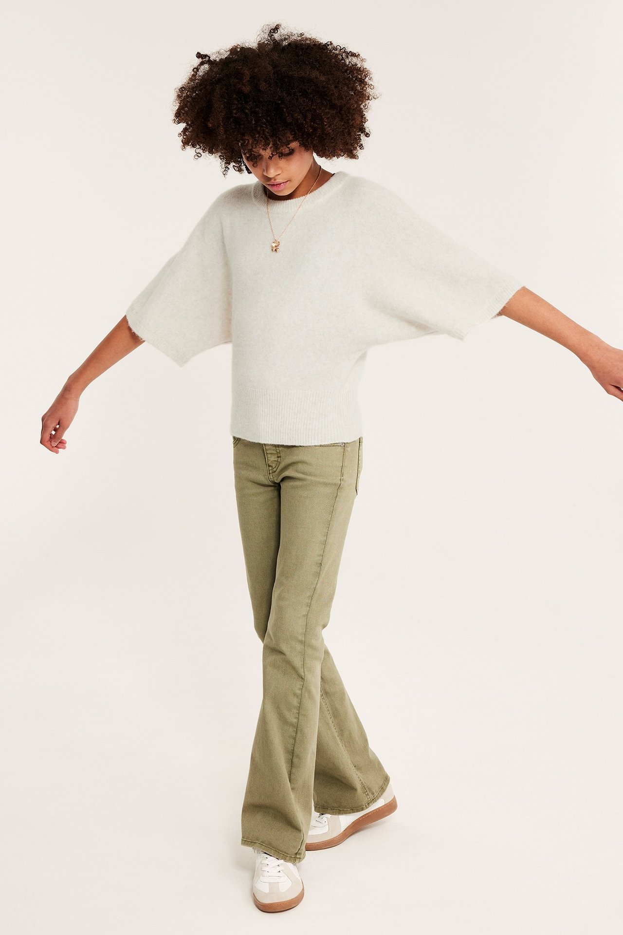 Frontvy av ett barn i melerad vit stickad topp med fladdermusärm, olivgröna utsvängda byxor och vita sneakers.