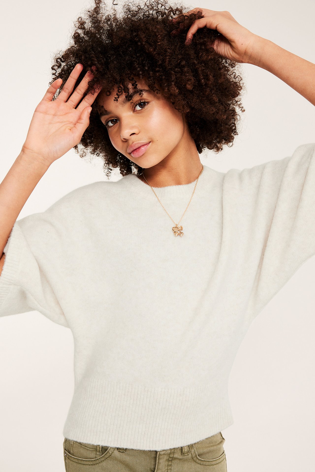 Framsida av kvinna i en lös, benvit melerad stickad topp, olivgröna byxor och ett guldhalsband.