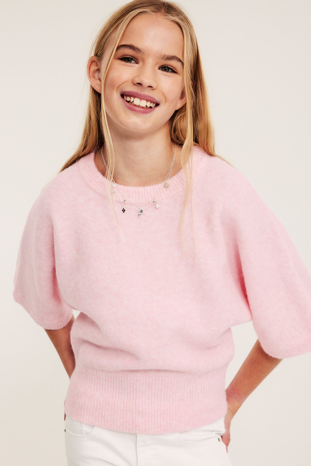 Frontutsikt: smilende barn i rosa strikket topp med vide ermer, hvite jeans og smykke med anheng.