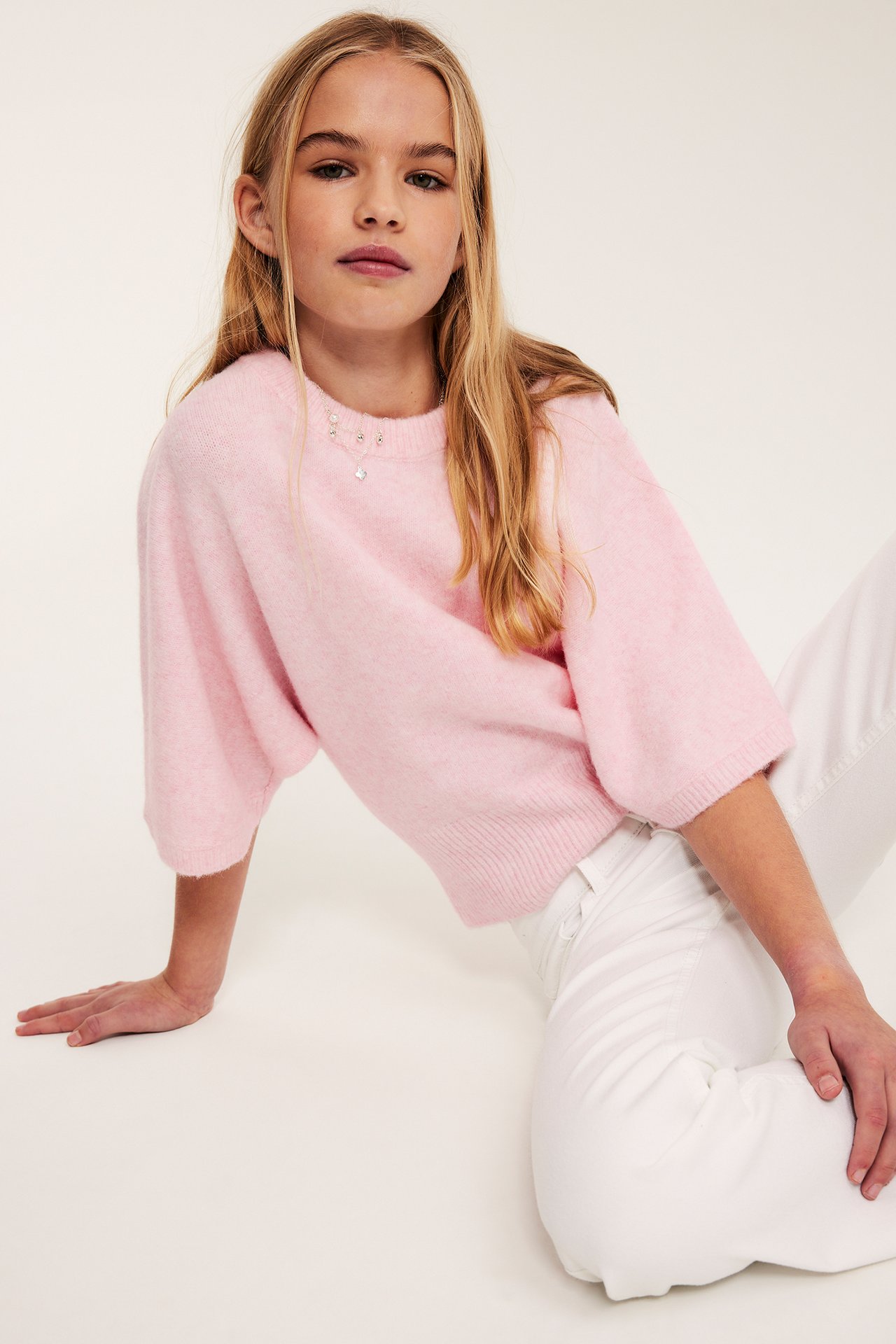 Lys rosa strikketop for barn med melert mønster, løs passform, over hvite bukser.