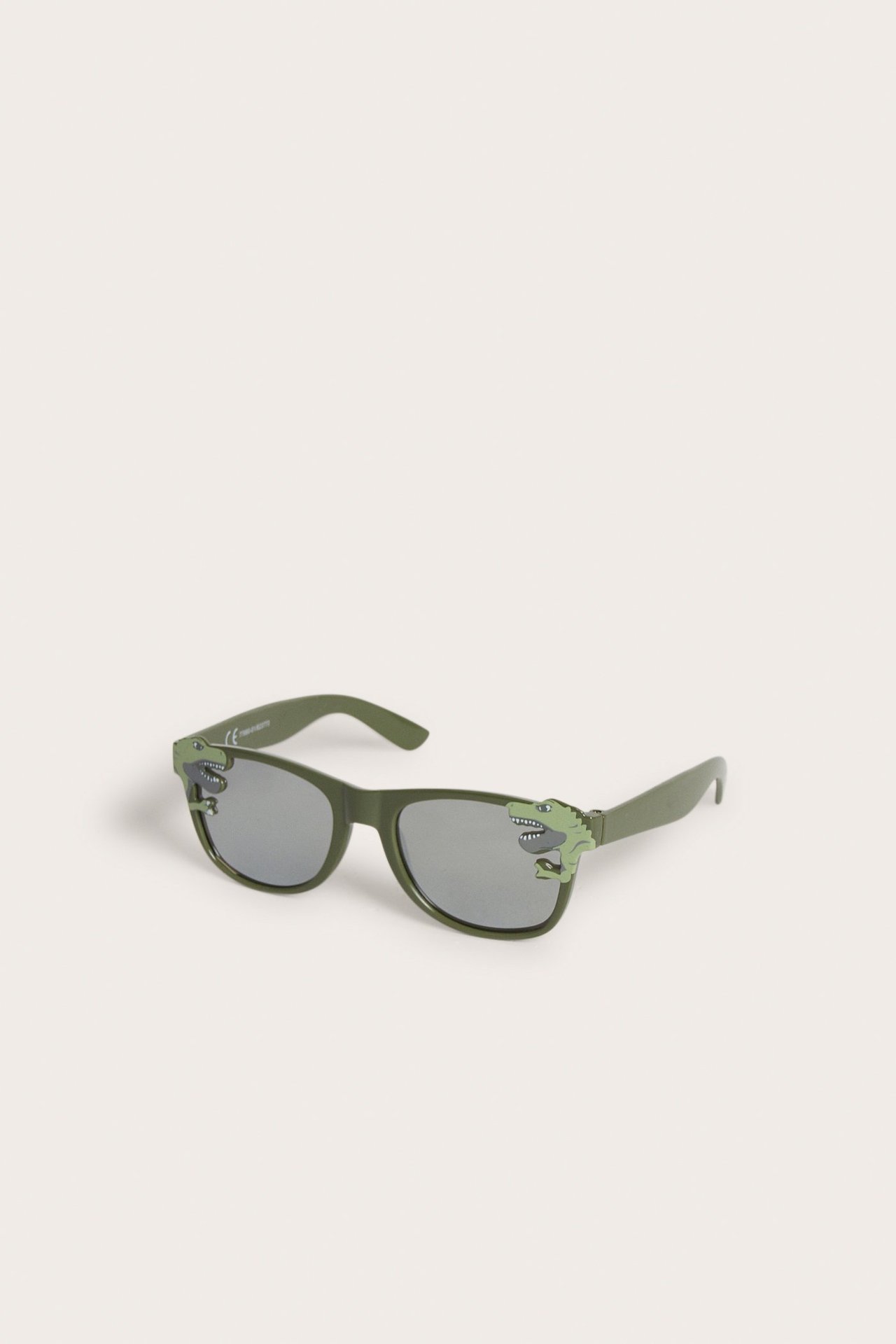 Viisto etunäkymä tummanvihreistä muovisista lasten aurinkolaseista, joissa on dinosauruskuvioita kehyksissä.
