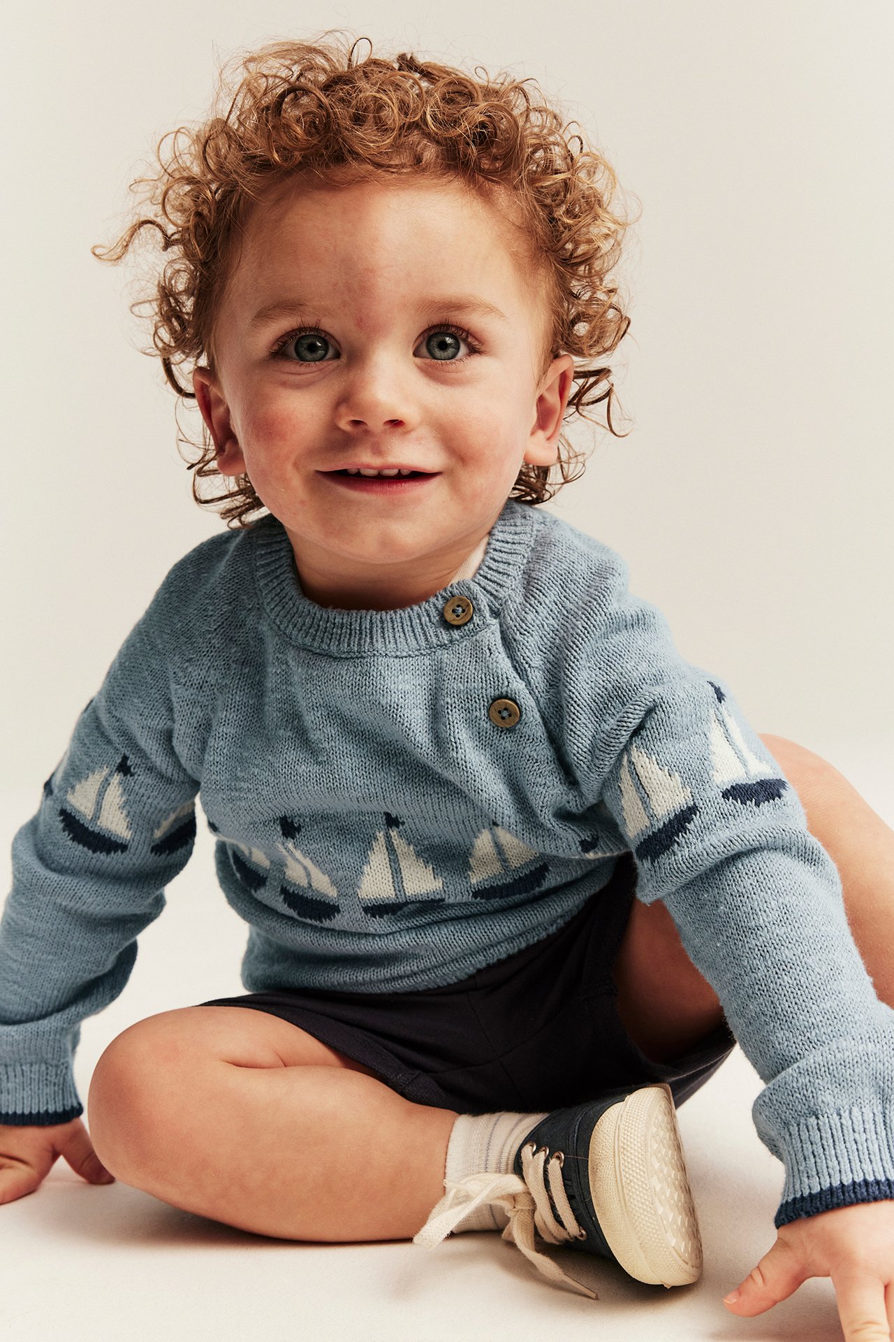 Edestä: vauva yllään vaaleansininen puuvillaneule purjevenekuvioilla, tummansiniset shortsit ja kengät.