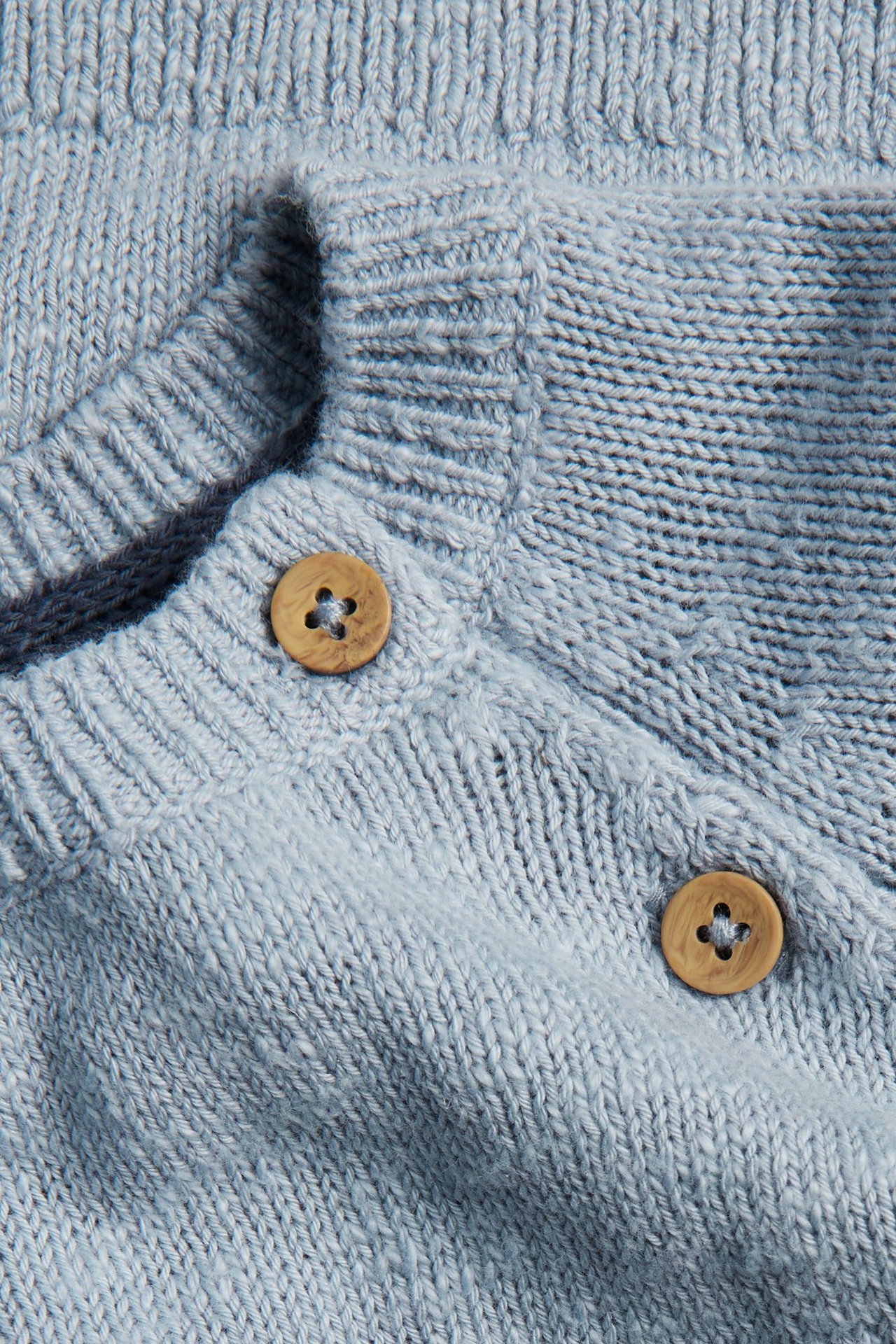Zbliżenie jasnoniebieskiego dzianinowego swetra z subtelnym wzorem żakardowym, ściągaczem i dwoma drewnianymi guzikami.