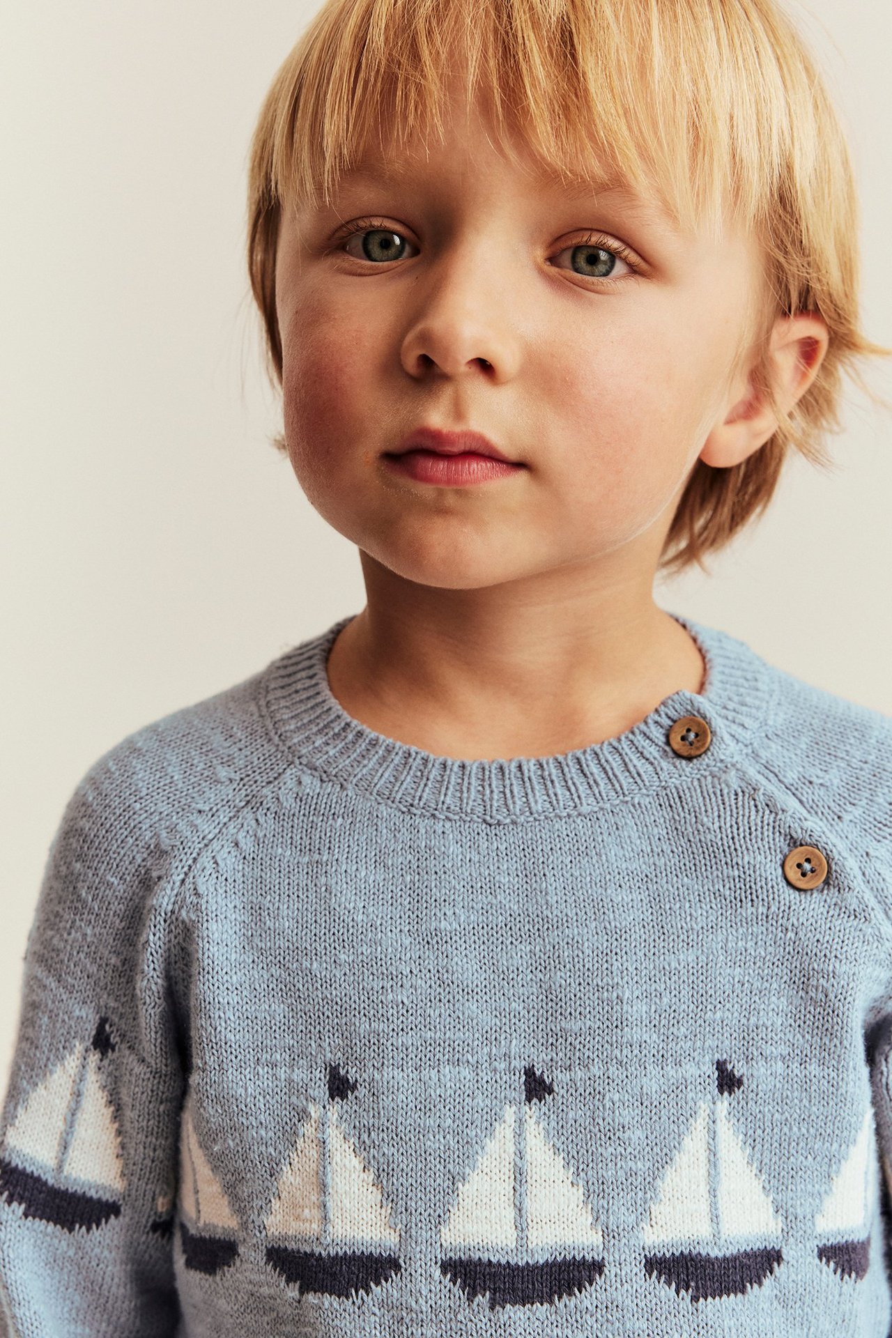 Barn i en lyseblå strikket bomullsgenser med hvitt seilbåtsjacquard og to skulderknapper.