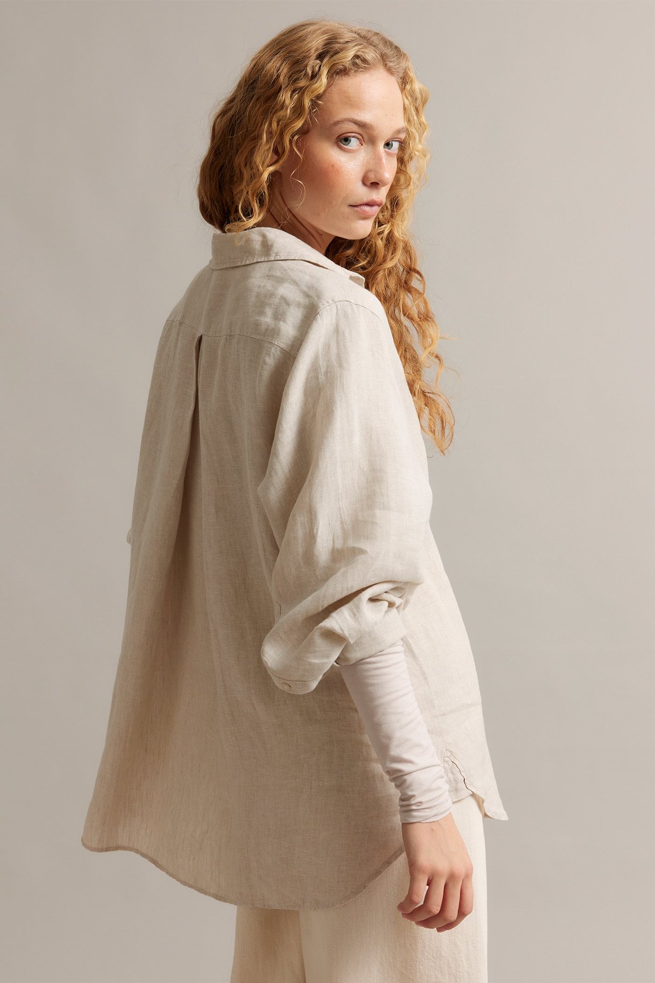 Kvinna i en oversized beige melerad linneskjorta med veck, buren över en beige topp och byxor.