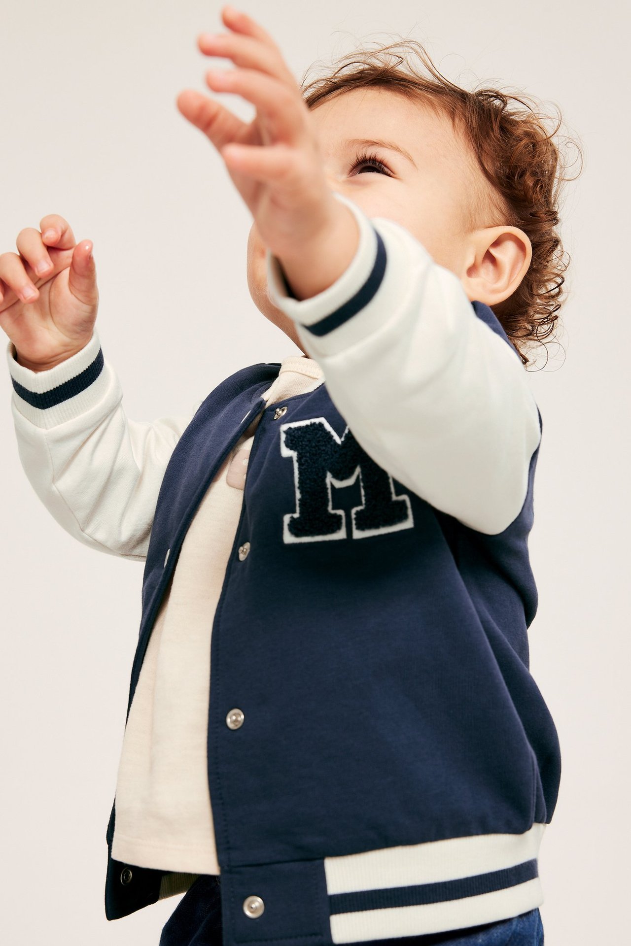 Edestä kuvattu: Vauva tummansinisessä baseball-takissa valkoisilla hihoilla, 'M'-applikaatio, ja vaalea t-paita.